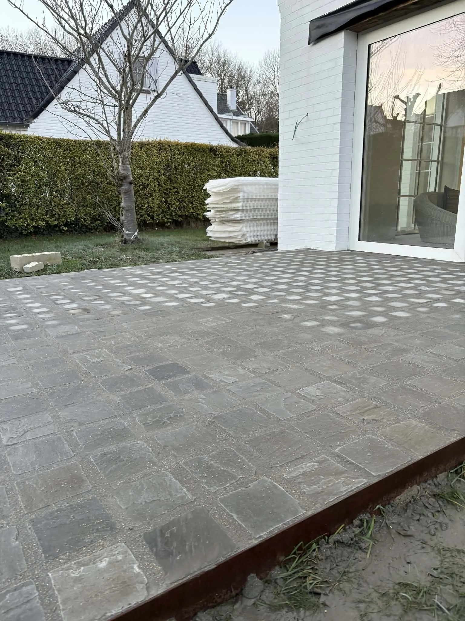 project: terras aanleg, locatie: Zuienkerke De afbeelding toont een tuin met een betegeld terras en een witte huismuur met grote glazen deur. Er staat een boom, een stapel witte isolatiepanelen en een groene heg op de achtergrond.