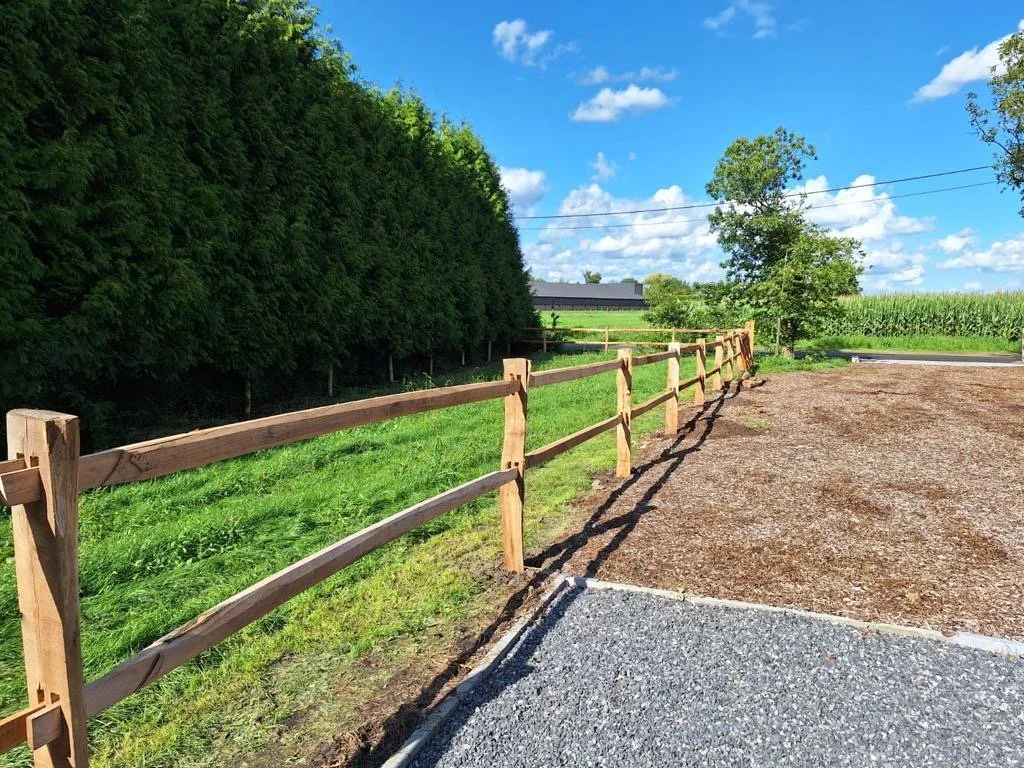 Een landelijke weg met houten hekwerk, groene bomen en een veld met maïs onder een blauwe lucht.