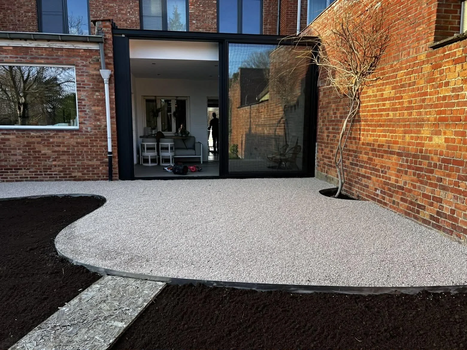 project: terras aanleg, locatie: Averbode nieuw aangelegd terras met een scheiding tussen donkere aarde en een lichtgrijze stenen of grind ondergrond, rondom een huis met een glazen schuifdeur, een boom zonder bladeren en een muur van rode bakstenen.