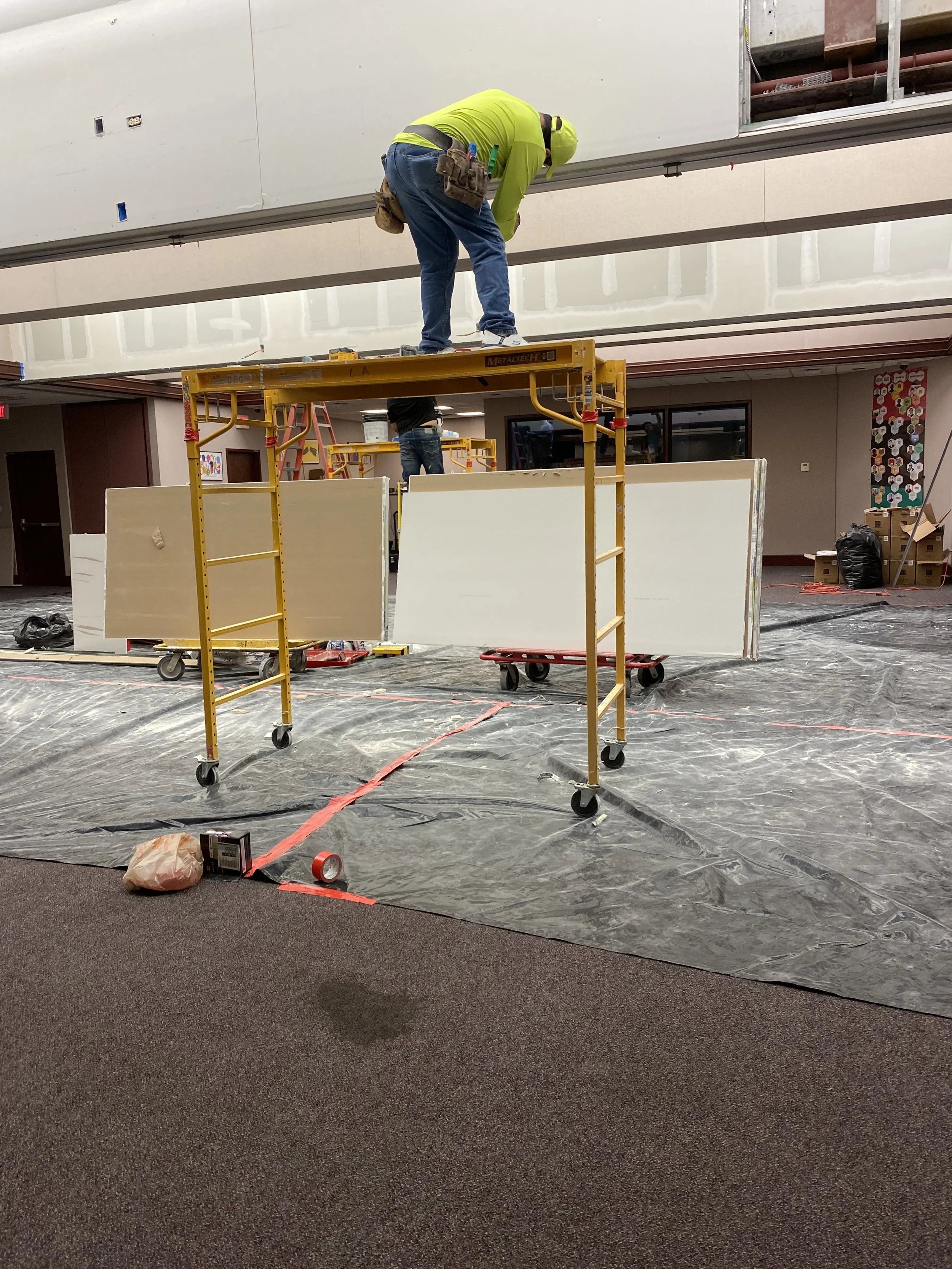 Construction workers installing or repairing drywall in an indoor space, with scaffolding and construction tools around.