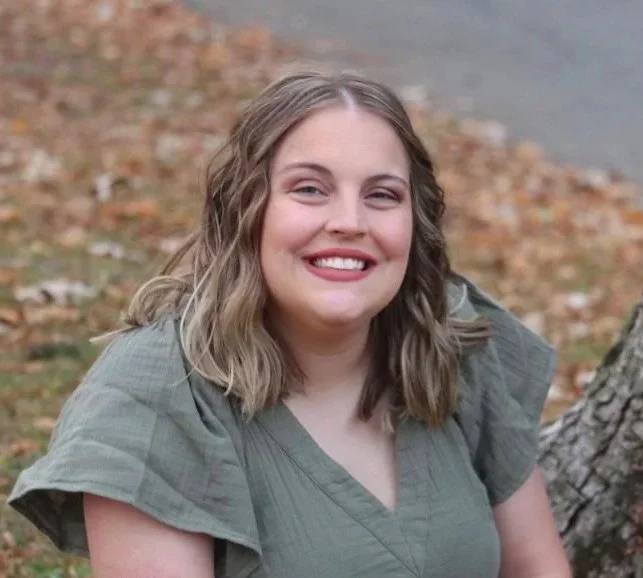Young woman with shoulder-length wavy blonde hair, smiling, outdoors in a park with fallen leaves and a tree trunk visible in the background.