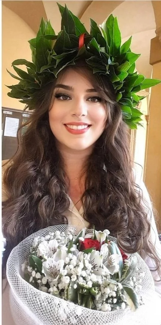 Una donna con lunghi capelli ricci, sorridente, mentre indossa una corona di foglie verdi e tiene in mano un bouquet di fiori bianchi con alcuni fiori rossi, il tutto in un ambiente interno. La Laurea di  Pianoforte Jazz di Oyku Dogan.