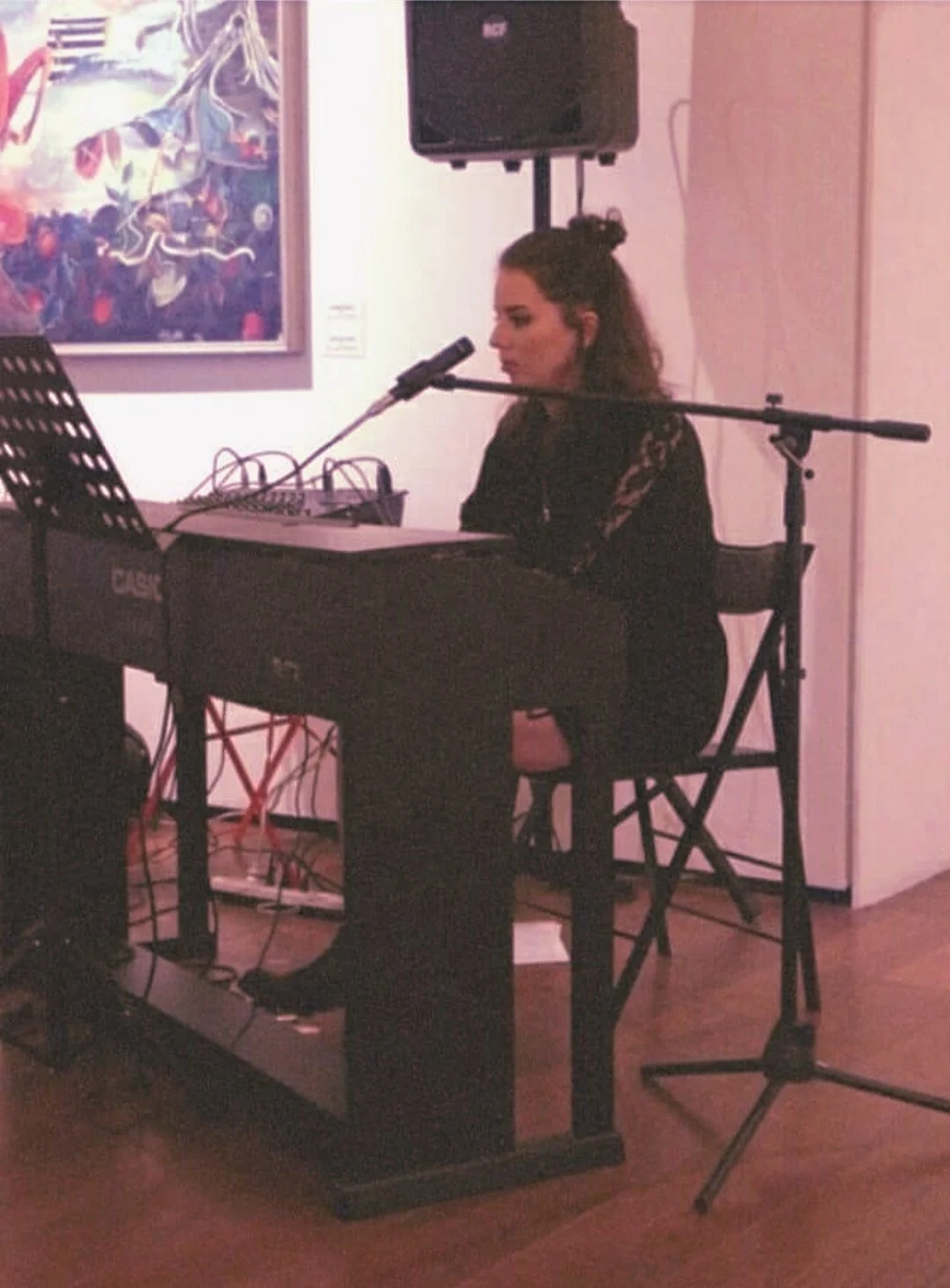 Donna che suona il pianoforte e canta davanti a un microfono in un ambiente artistico, con un quadro colorato appeso alla parete