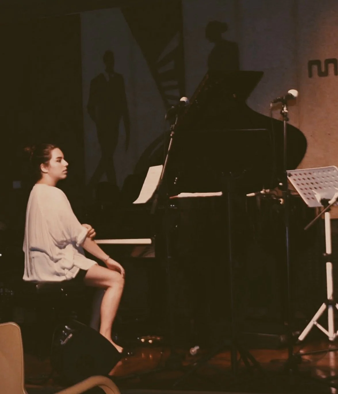 Una giovane donna seduta al pianoforte durante una performance musicale, con una stanza scura e un microfono davanti a lei.