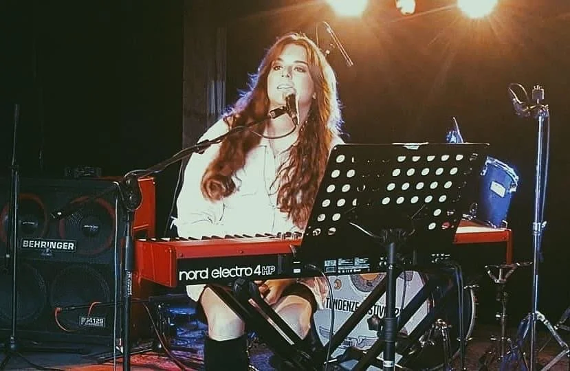 Una donna con lunghi capelli ricci canta e suona un sintetizzatore rosso sul palco in un locale di musica con luci calde.