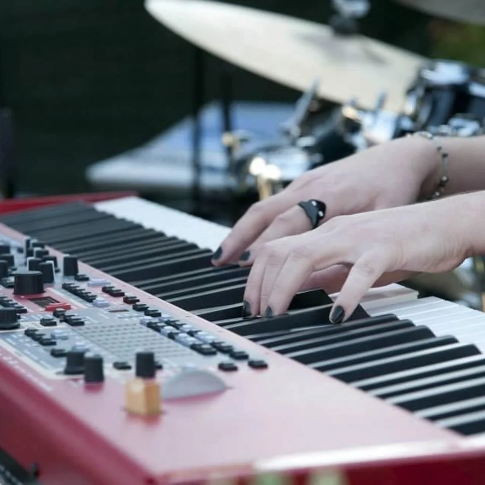 Mani che suonano un sintetizzatore musicale con tasti neri e bianchi, con uno sfondo di strumenti musicali.