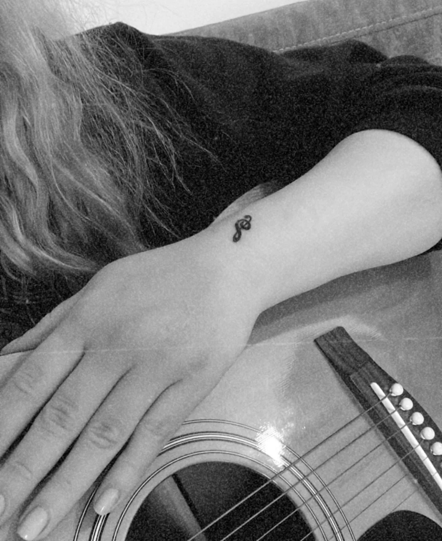 Mano di una persona con tatuaggio a forma di nota musicale sulla parte superiore del braccio, che poggia su una chitarra acustica. La persona ha i capelli lunghi e ricci. Immagine in bianco e nero.
