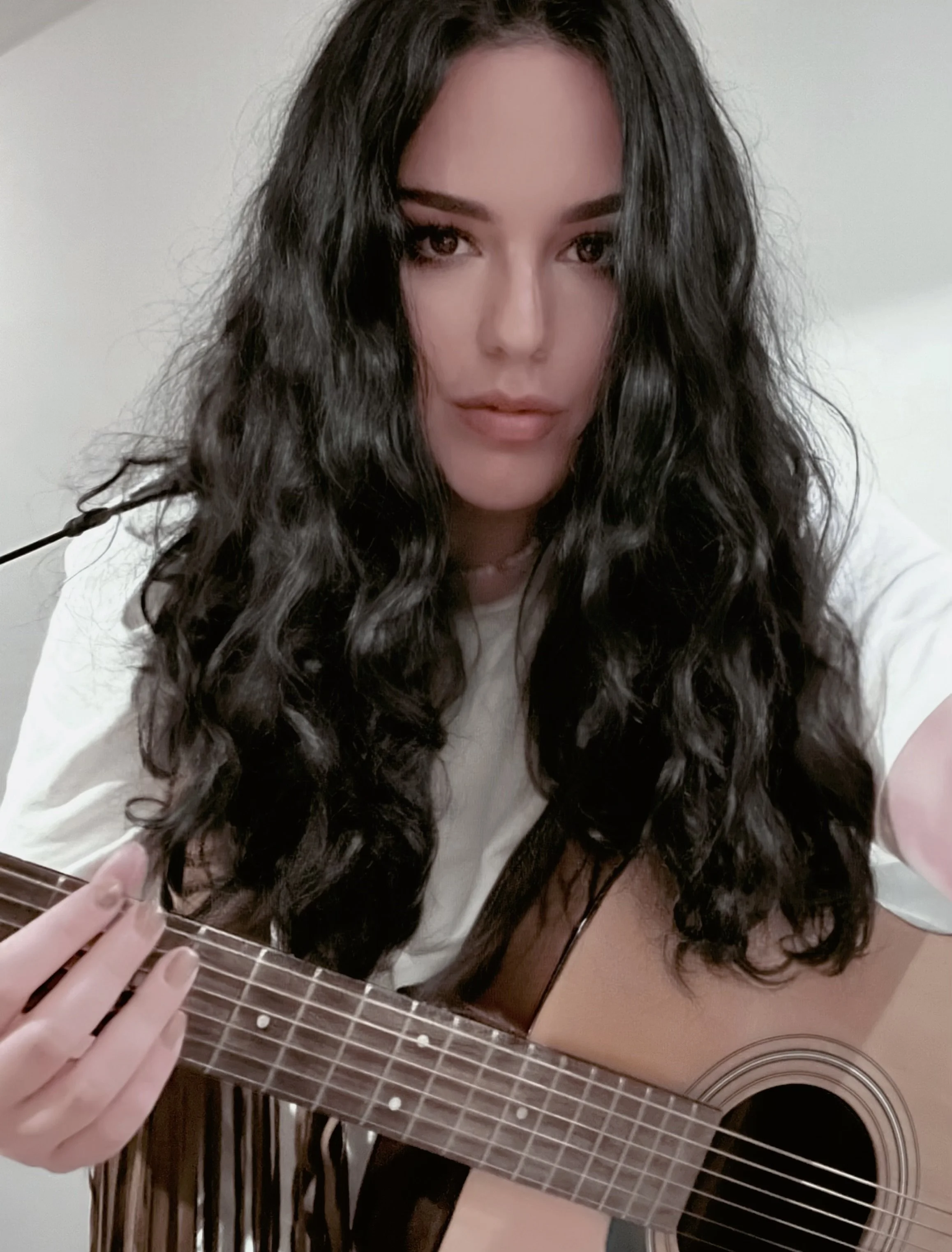 Una donna con capelli ricci neri su uno sfondo chiaro, seduta con una chitarra acustica, che posa per una foto.