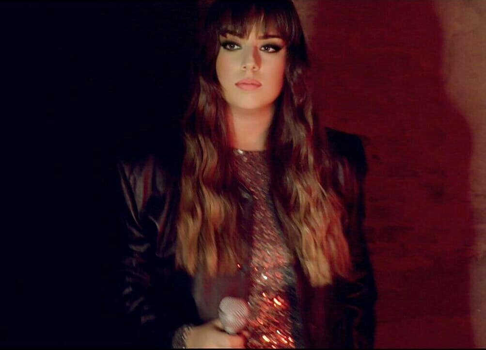 Donna con capelli lunghi, ricci e biondi, trucco intenso, indossa una giacca nera e una maglietta con paillettes, tiene un microfono in mano, sfondo scuro.