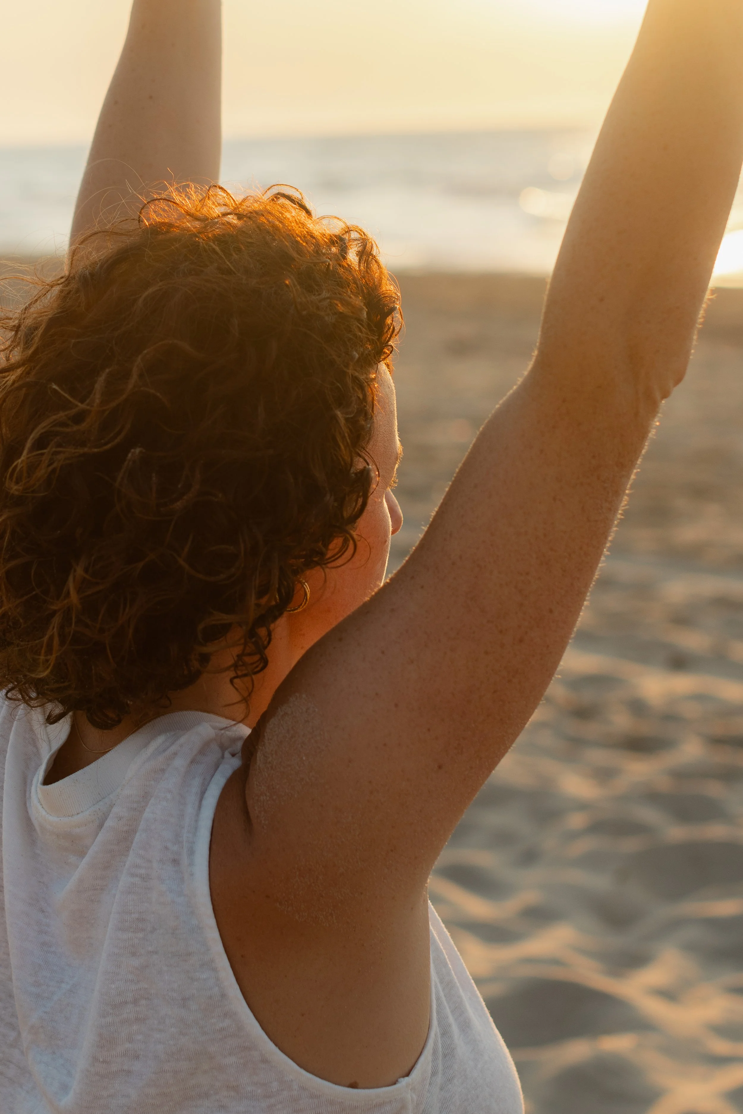 A person with curly red hair raising her arms on the beach during sunset, wearing a white sleeveless top.