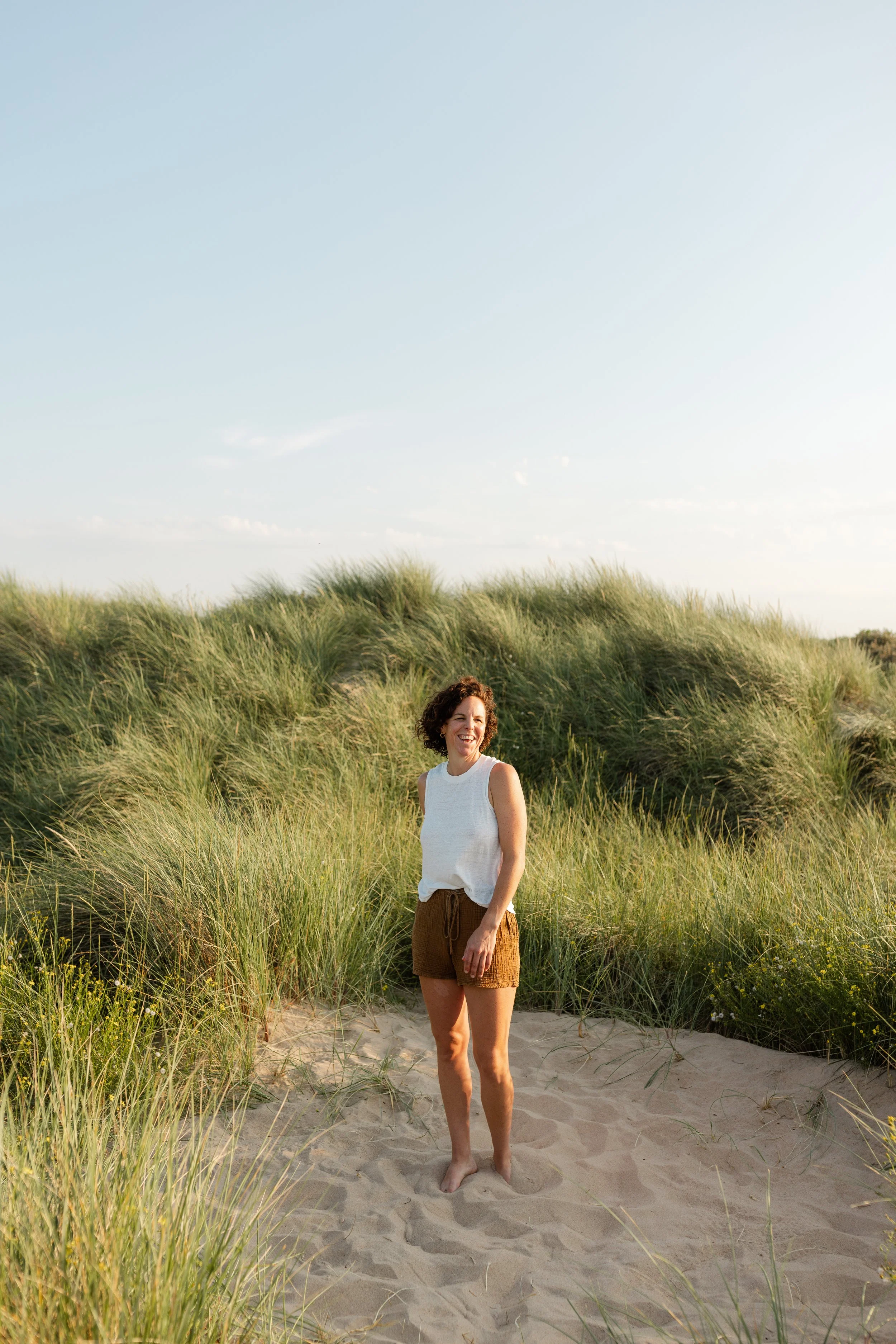 A woman with curly hair wearing a white sleeveless top and brown shorts, standing barefoot on a sandy path amidst tall green beach grass, smiling outdoors with a clear sky overhead.