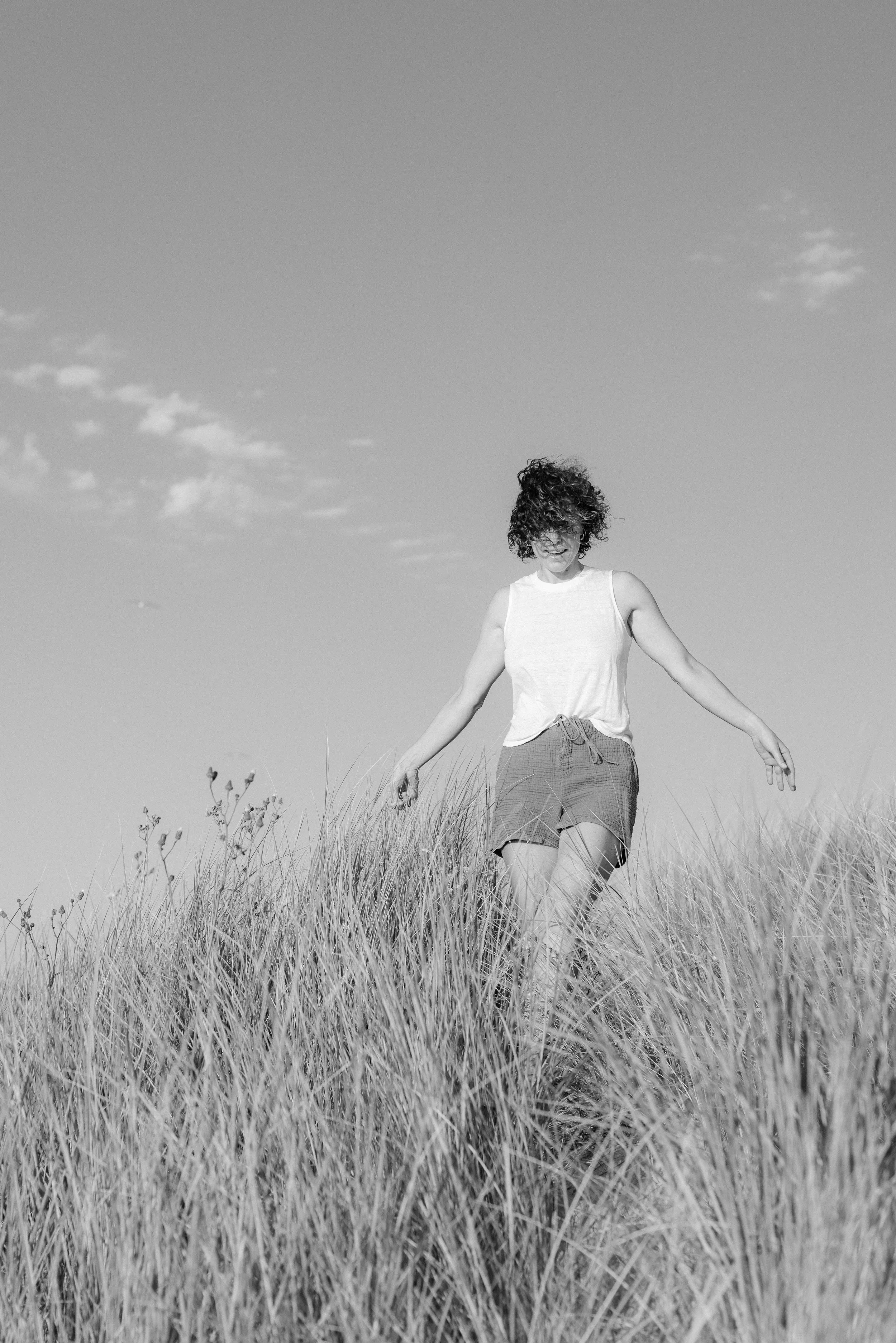 Black and white photo of a smiling woman with curly hair wearing a sleeveless top and shorts, walking through tall grass outdoors, under a partly cloudy sky.