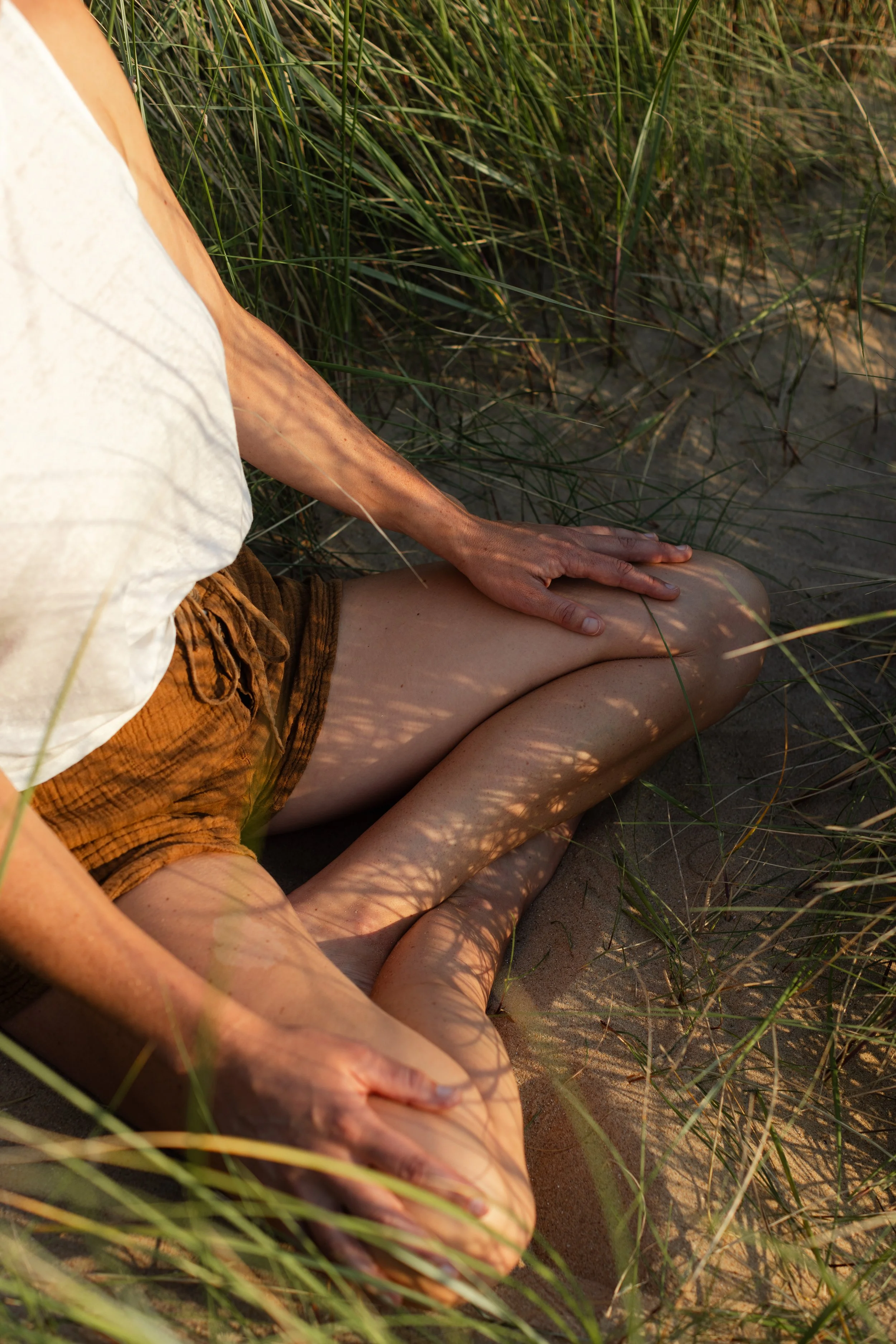 A person sitting on sandy ground with legs crossed, wearing orange shorts and a white shirt, surrounded by tall green grass.