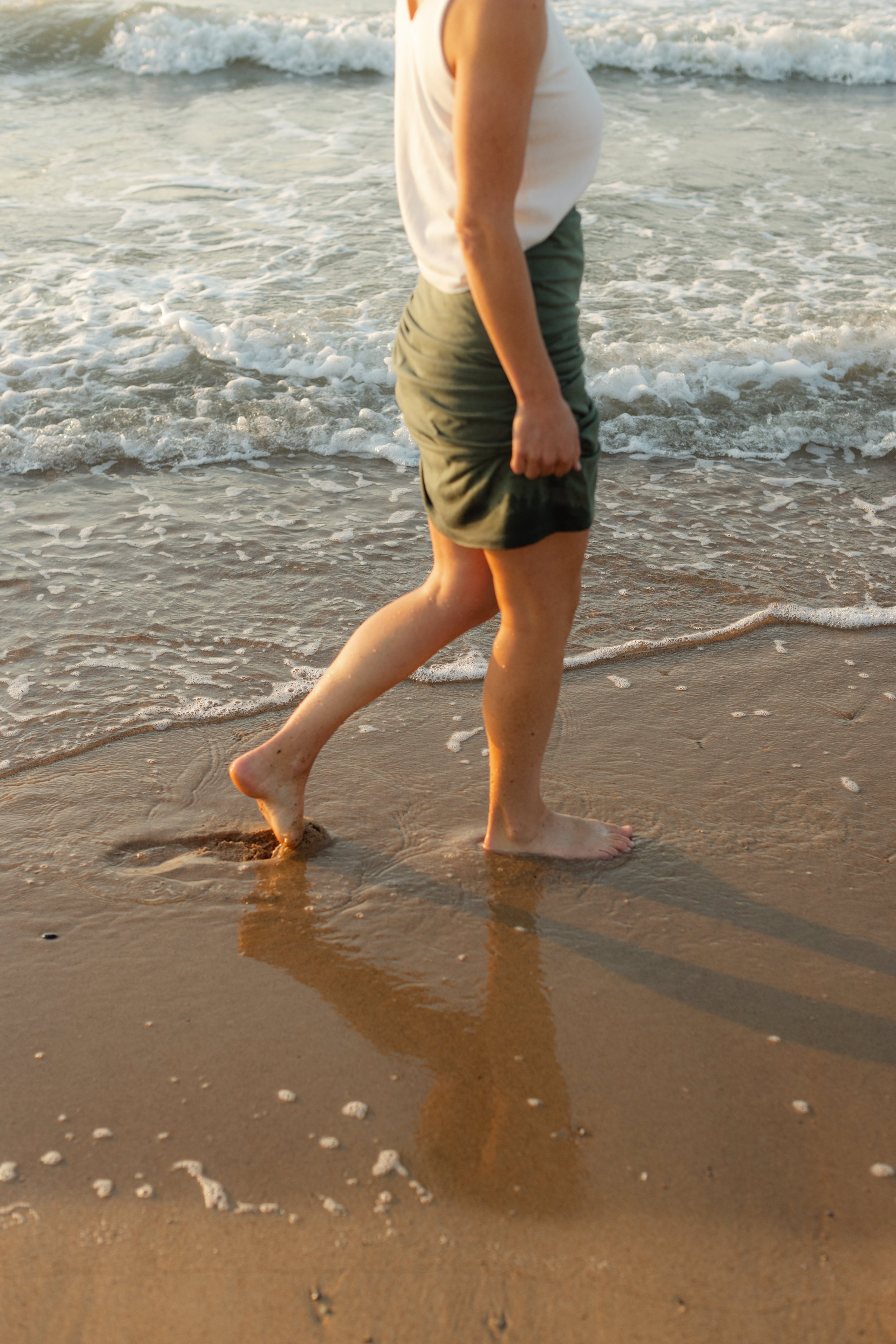 Partial view of a person walking barefoot on the wet sand at the beach with ocean waves in the background, during sunset.
