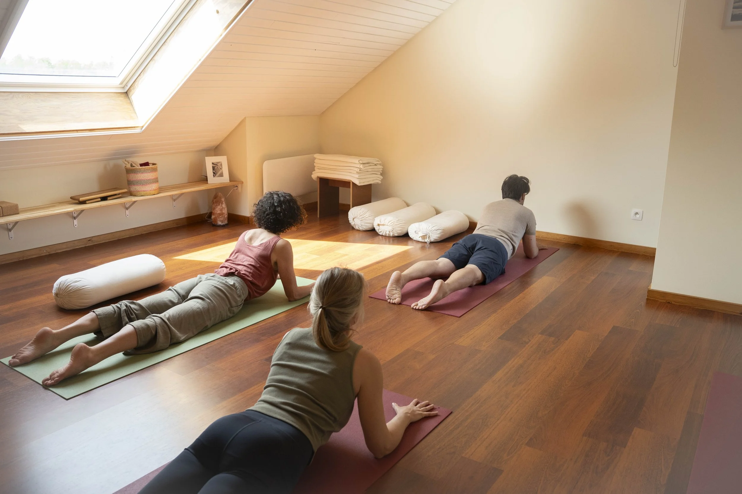 People practicing yoga in a bright attic room with sloped ceiling, sunlight through a skylight, wooden floor, and minimal decor.