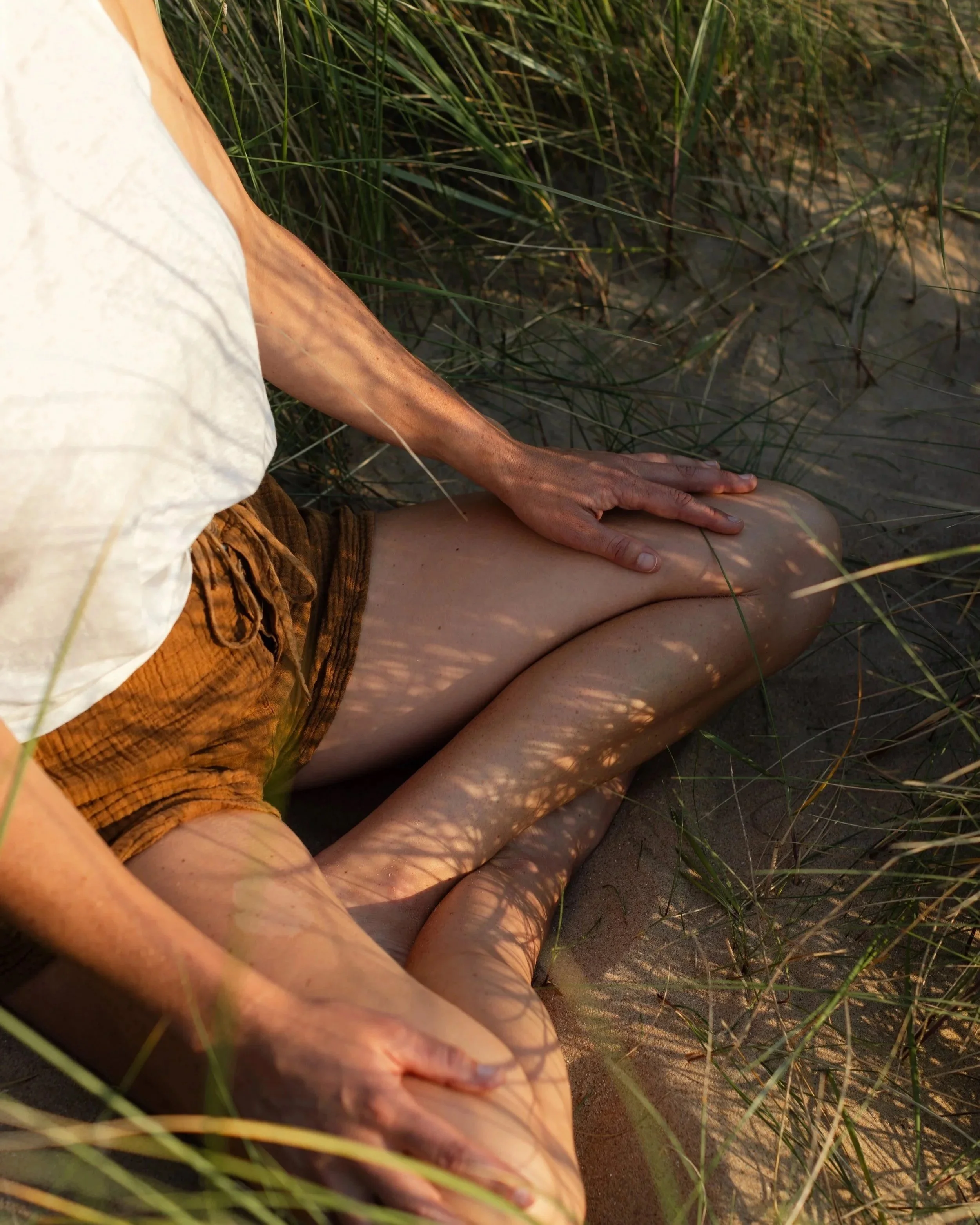 Person sitting on sandy ground with grass surrounding, wearing a light-colored top and patterned shorts.