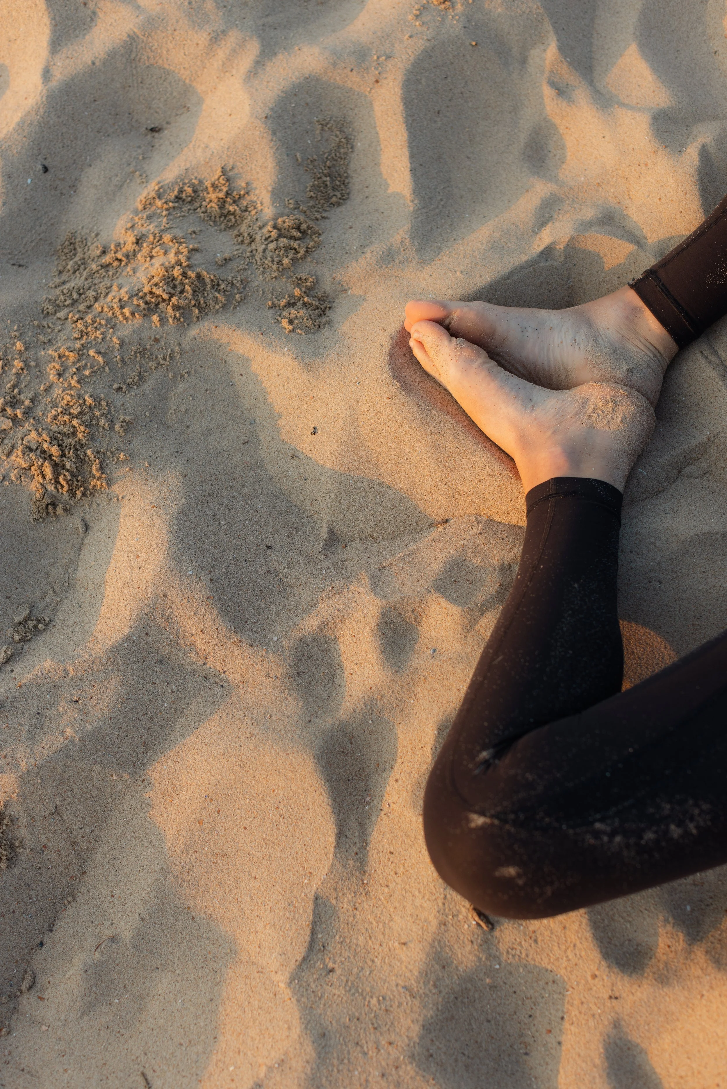 A person's hand and leg sandy beach, with the person wearing a black wetsuit, and the hand making a flat gesture on the sand that appears to be a fish.