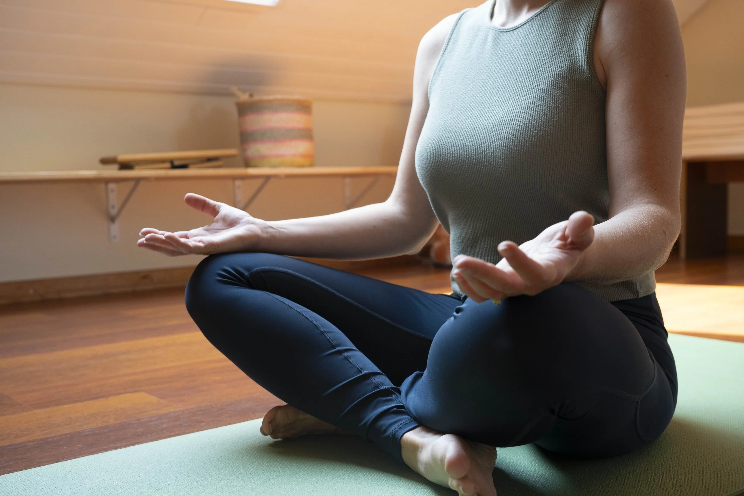 A person practicing yoga in a kneeling cross-legged position, with hands resting on their knees, in a room with wooden flooring and a basket in the background.