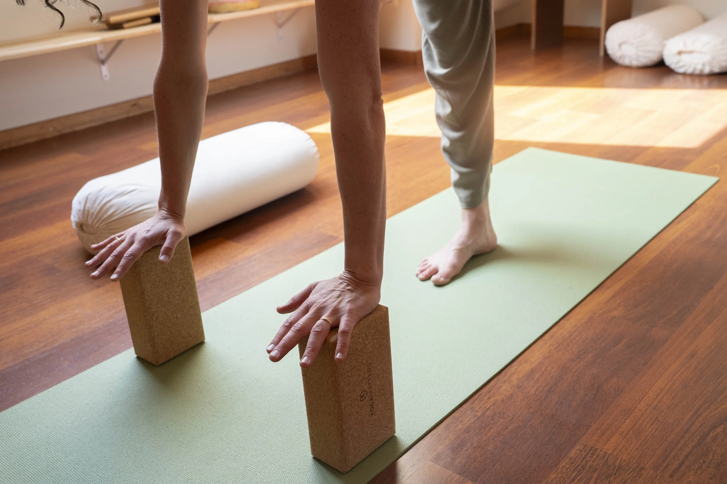 A person practicing yoga in a studio, using cork blocks to support their hands while in a downward dog pose on a green yoga mat.