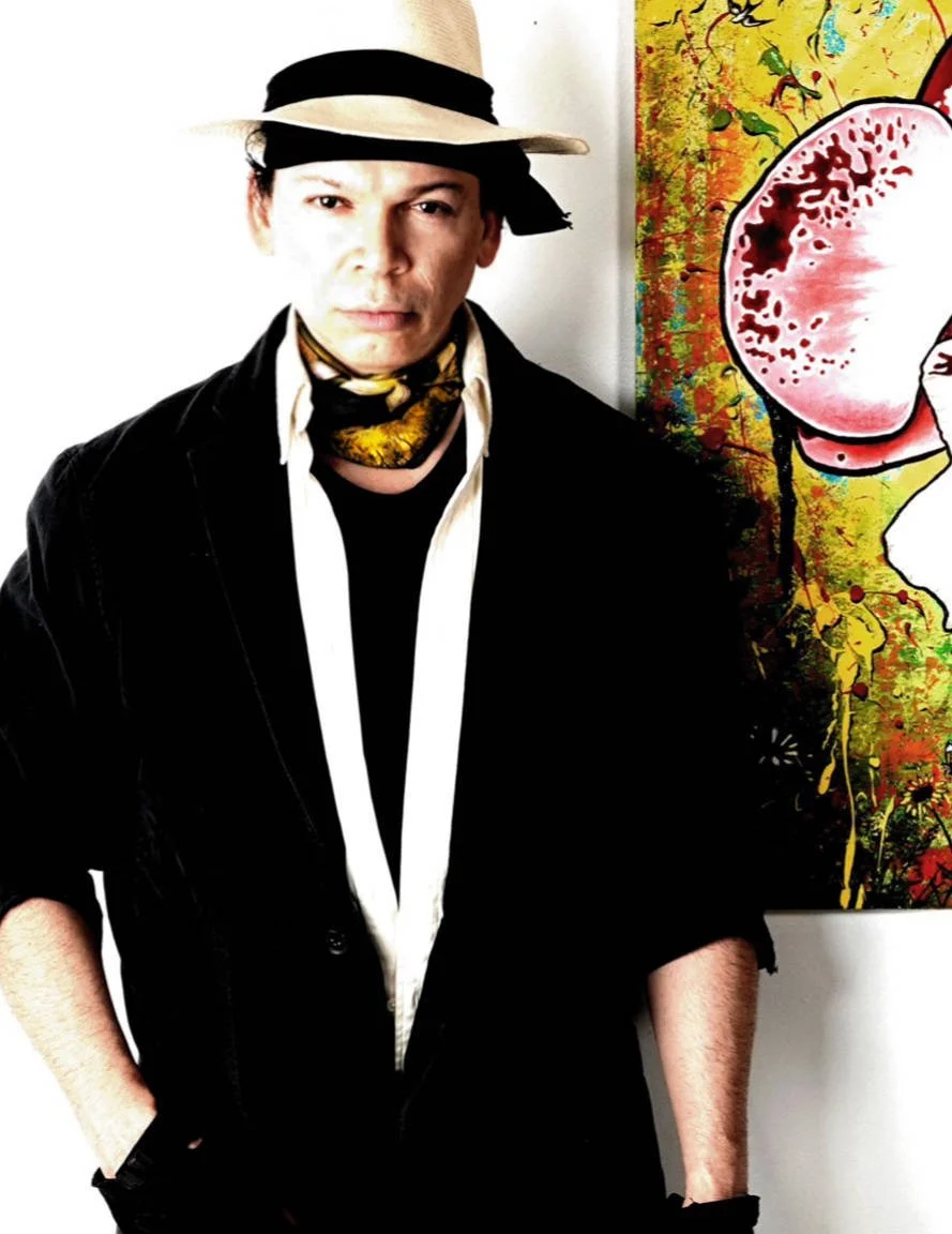 Von Taylor wearing a light-colored fedora with a black band, black jacket, white shirt, and a patterned scarf standing in front of an abstract colorful painting.