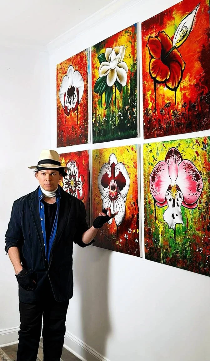 Von Taylor wearing a dark jacket, hat, and gloves standing in an art gallery, pointing at colorful paintings of flowers on the wall.