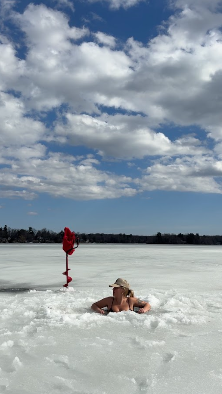 Woman swimming in a frozen lake in winter, wearing a hat, with a red bag hanging on a stick nearby, under a partly cloudy sky.