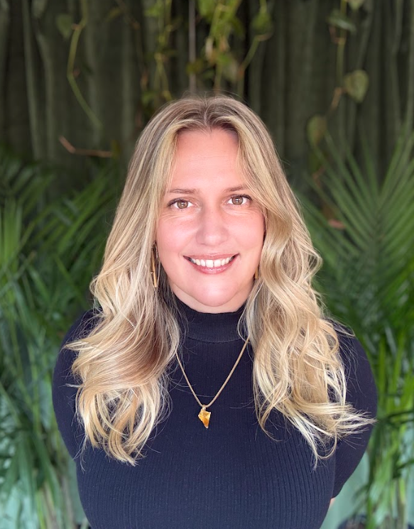 A woman with blonde wavy hair, smiling, wearing a black turtleneck sweater, gold necklace, and gold earrings, standing in front of green tropical plants.