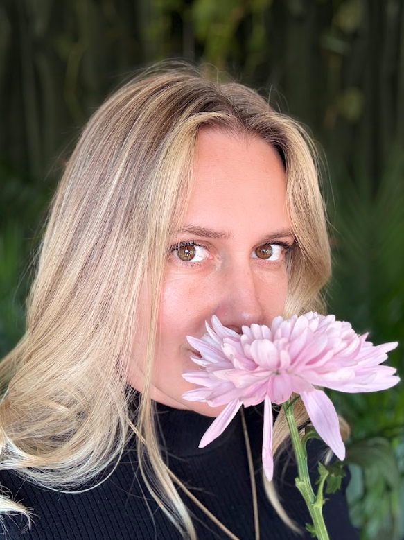 A woman with blonde hair holding a pink chrysanthemum flower close to her face in front of green foliage.