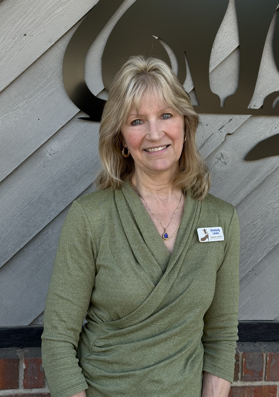 A woman with blonde hair, wearing a green top, standing in front of a wooden wall with a decorative black wrought iron element. She is smiling and wearing a name tag that reads 'Kimberly Lewis' with a logo of a kangaroo.