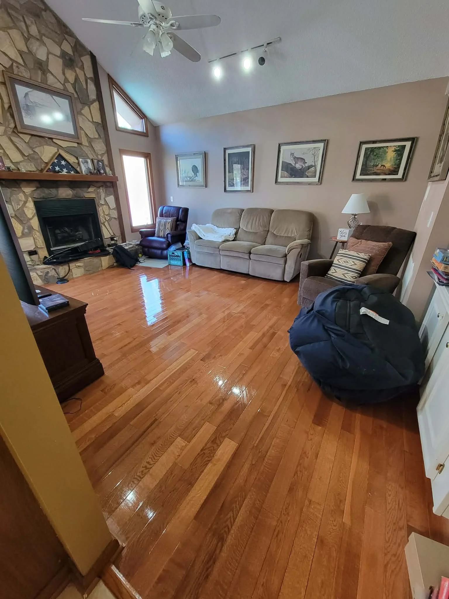 A living room with hardwood floors, a stone fireplace, and various framed pictures on the wall. There are three chairs, including a beige couch, a dark brown armchair with a lamp, and a purple recliner near a window. Items such as a black backpack, a