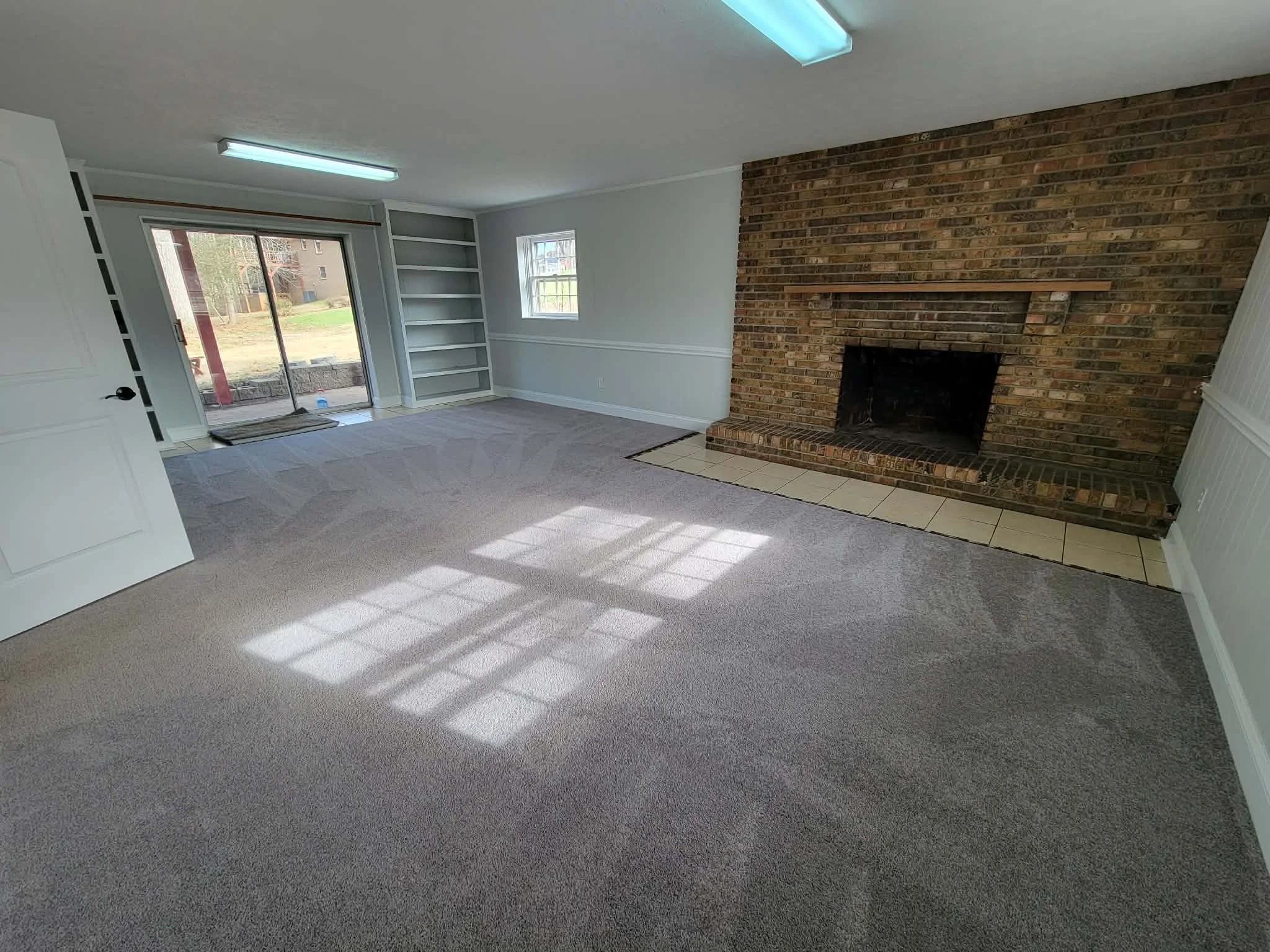 Empty living room with gray carpet, brick fireplace, built-in white bookshelves, sliding glass door leading outside, and sunlight coming through the window.