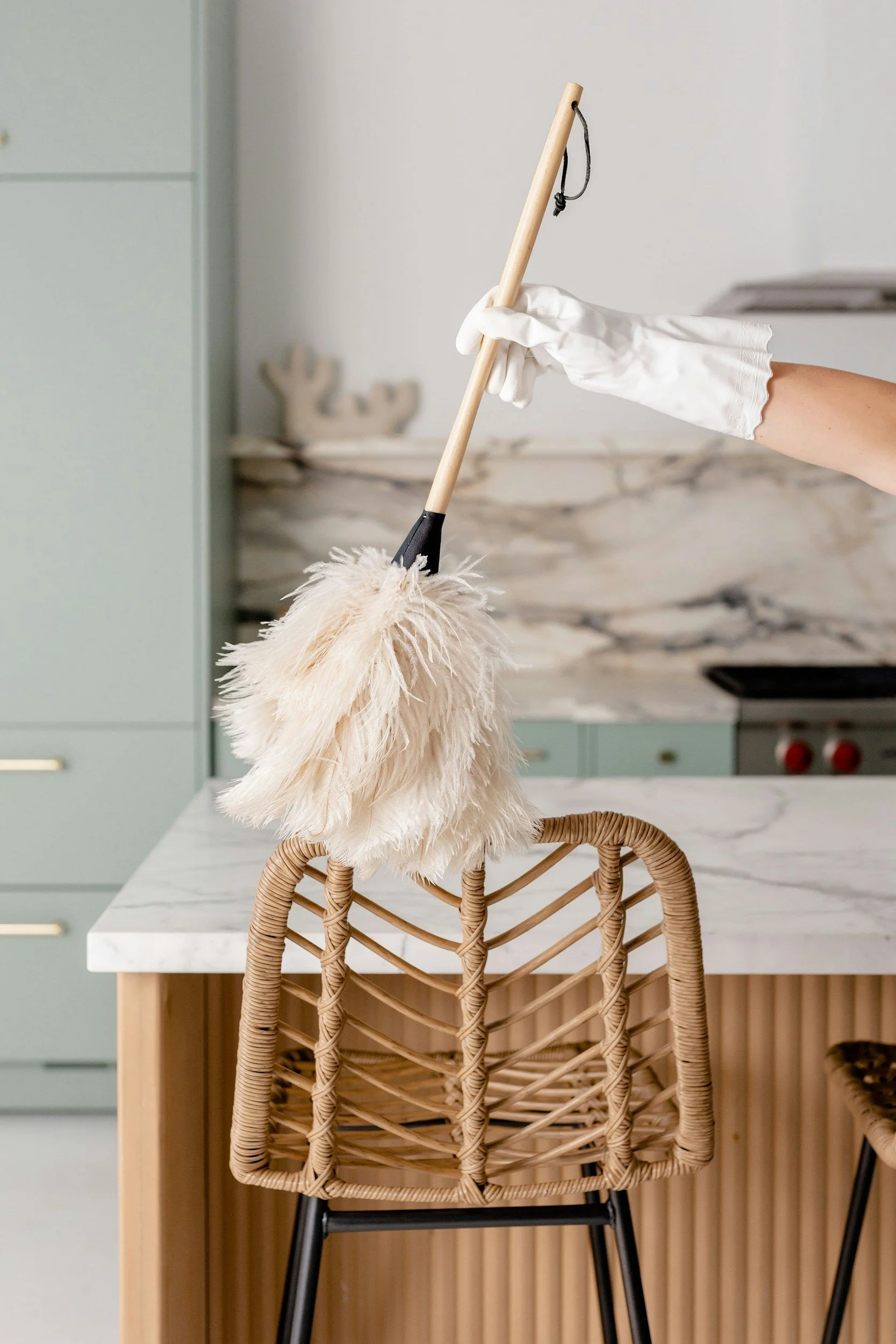 A white feather duster hanging from a long-handled tool, held by a person wearing white gloves, with a kitchen background featuring marble countertops and light-colored cabinetry.