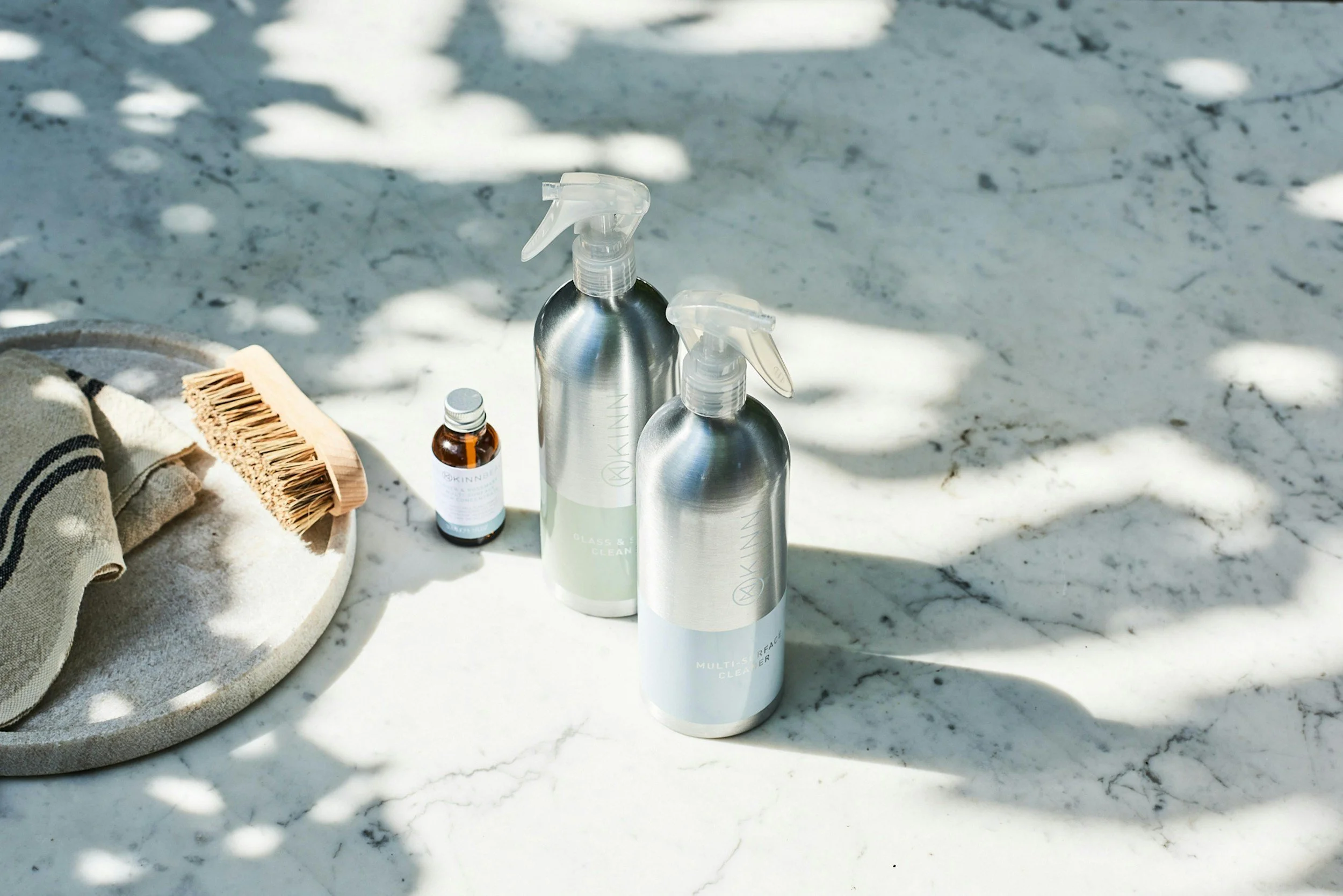 Two metallic spray bottles, a small brown vial, a wooden brush, and a rolled towel on a marble surface, with shadows of leaves overhead.