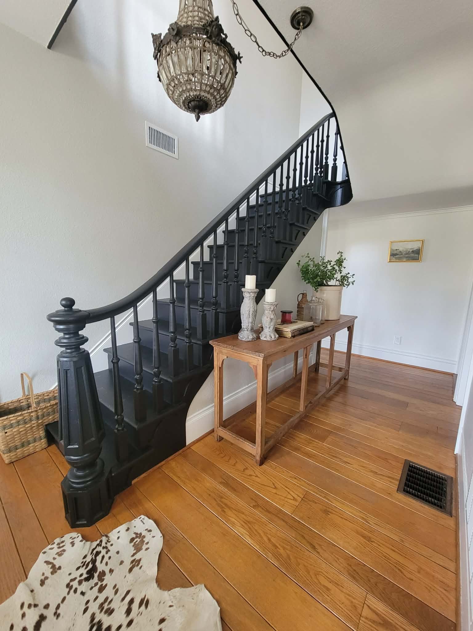 Interior of a house showing a curved staircase with black banisters, a hanging chandelier, a wooden table with decorative items, plant, candles, and framed art on the wall.