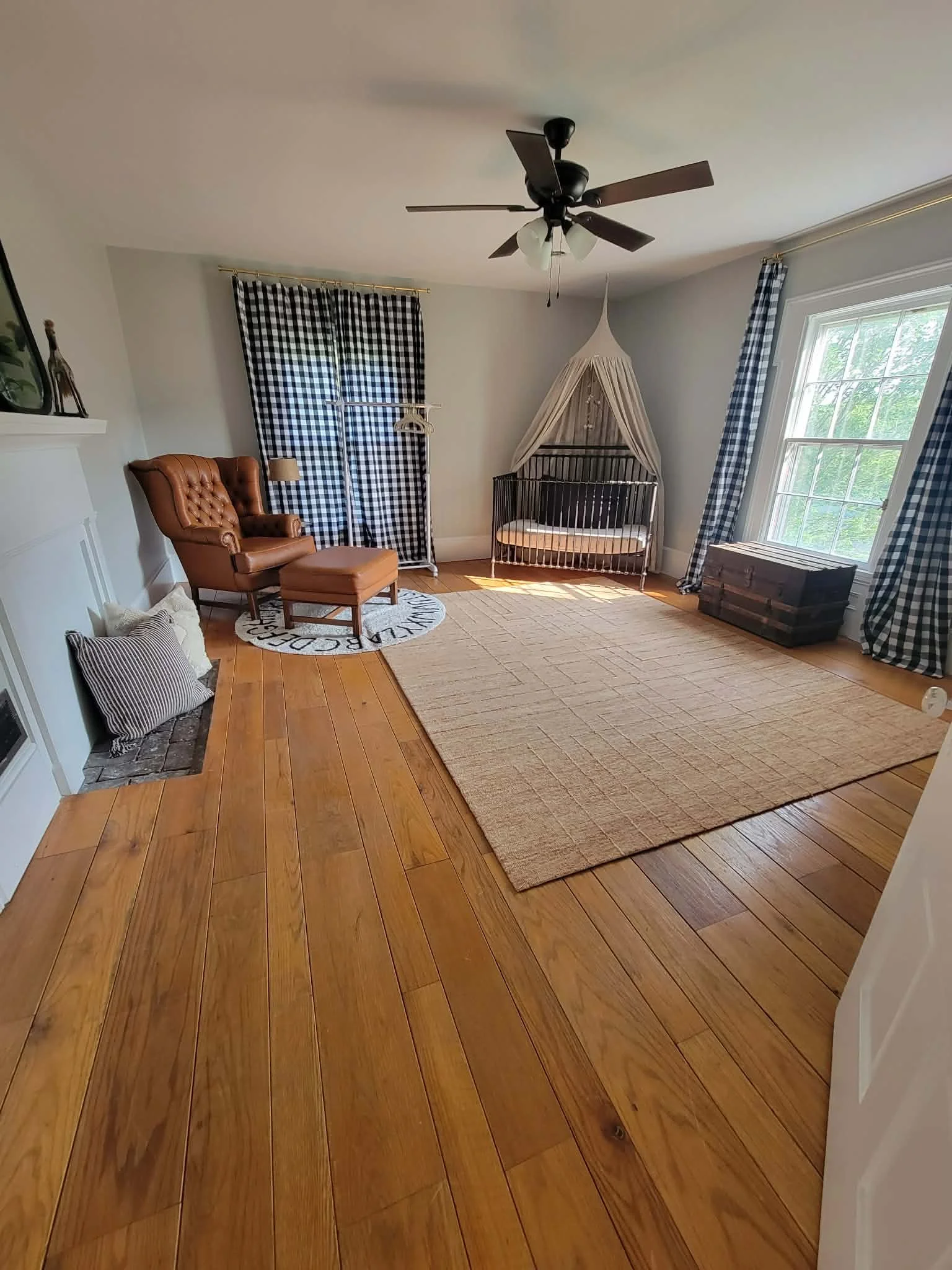 A bright living room with hardwood floors, a large beige area rug, a black ceiling fan, a white canopy crib in the corner, a brown tufted armchair with an ottoman, plaid curtains, a small round rug with text, a wooden trunk near the window, and a fir