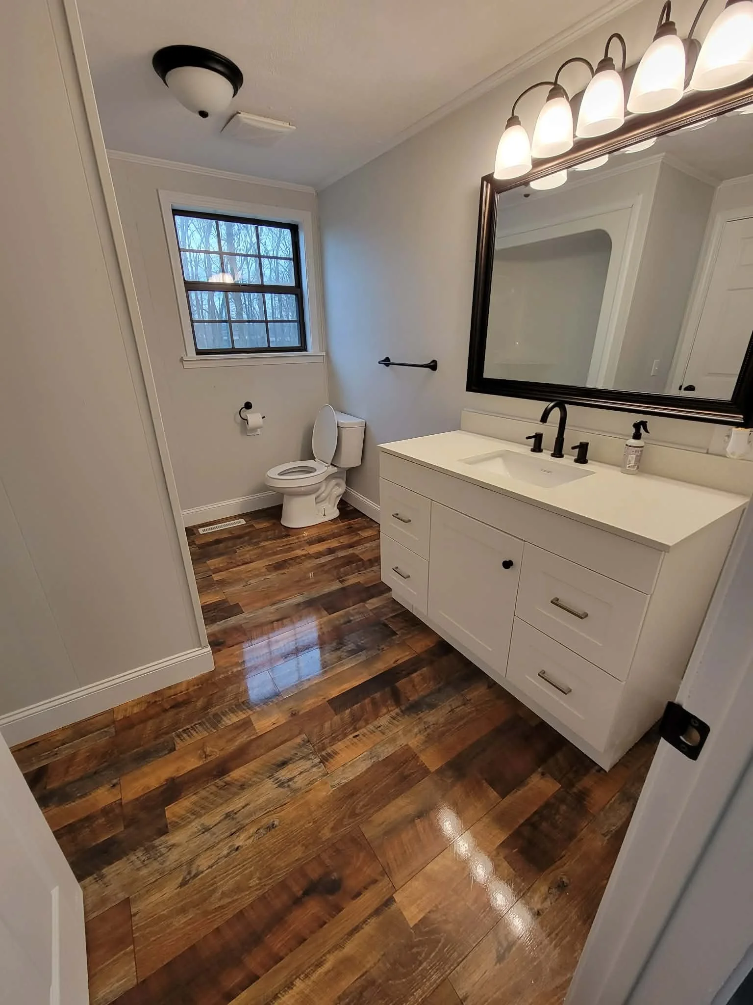 A small bathroom with a white vanity, black faucet, large wall mirror, and a row of four lights above it. There is a toilet with a black toilet paper holder on a light gray wall, a window with a view of tree branches outside, a black grab bar, a ceil