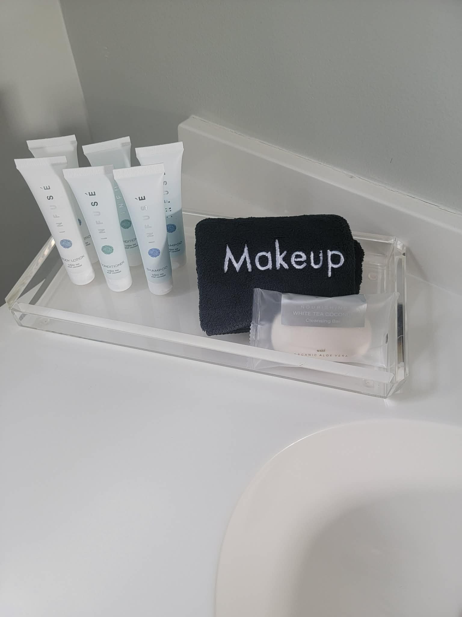 Travel-sized toiletries including shampoo, conditioner, body lotion, and soap, with a black washcloth labeled 'Makeup' and a small sealed packet, on a clear acrylic tray beside a sink.