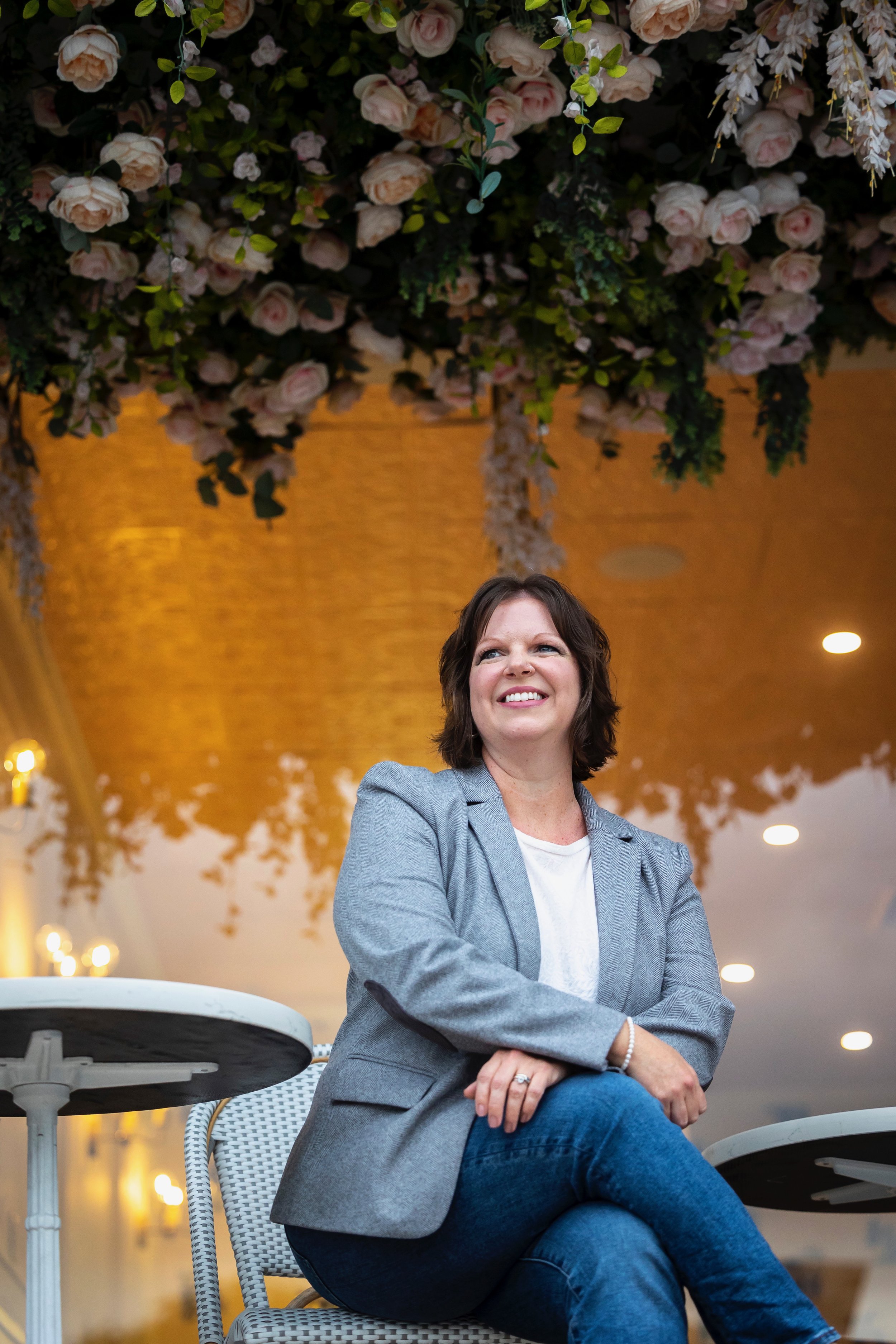 A woman sitting at an outdoor cafe, smiling, with a large ceiling installation of pink and white roses and greenery overhead, and warm lighting in the background.