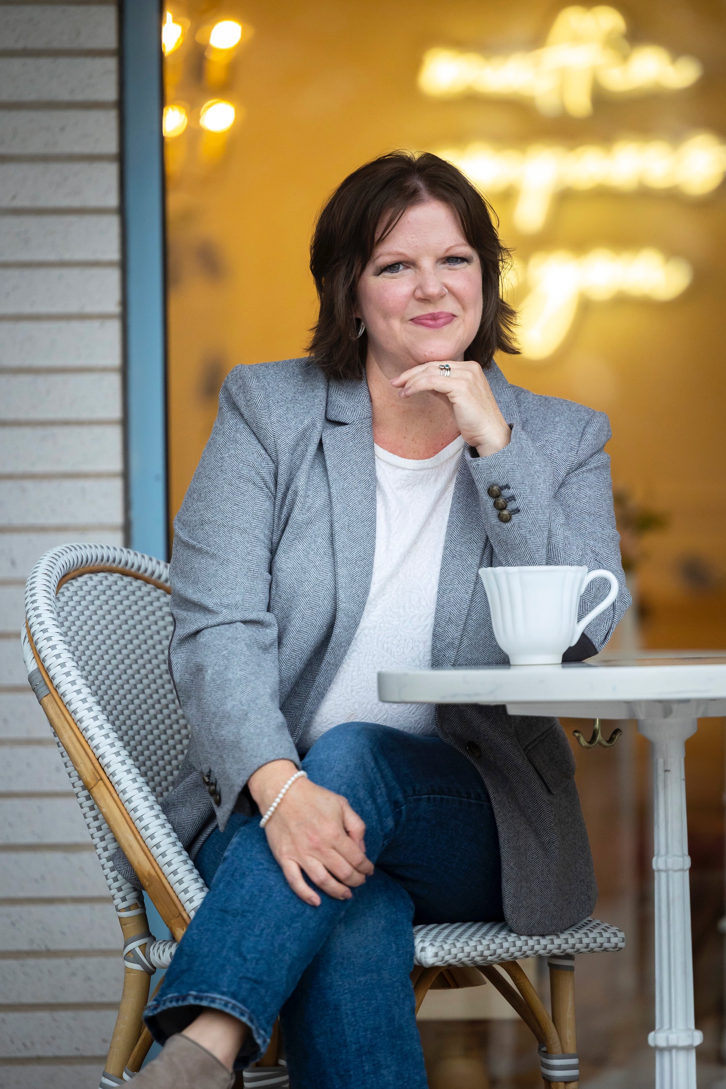A woman sitting outdoors at a cafe table with a coffee mug in front of her, wearing a gray blazer and white shirt, with short brown hair, smiling and looking at the camera.