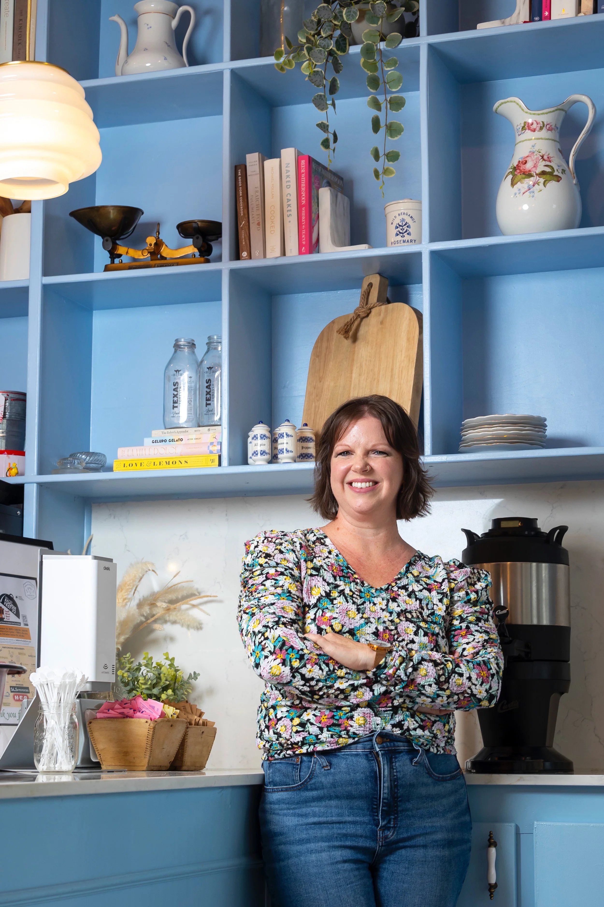 A woman with short brown hair smiling and crossing her arms in a kitchen or café setting, with blue shelves full of books, vases, and decorative items behind her, and a coffee machine on the counter to her right.