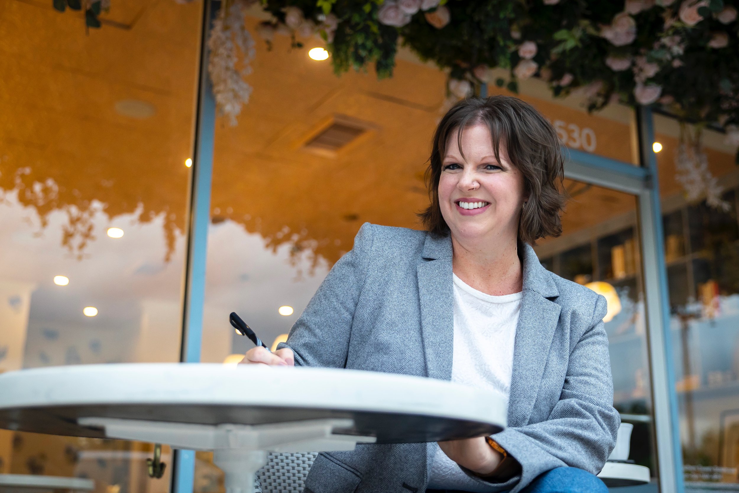 A woman smiling and writing on a notepad at an outdoor café with a glass storefront and trees reflected in the background.