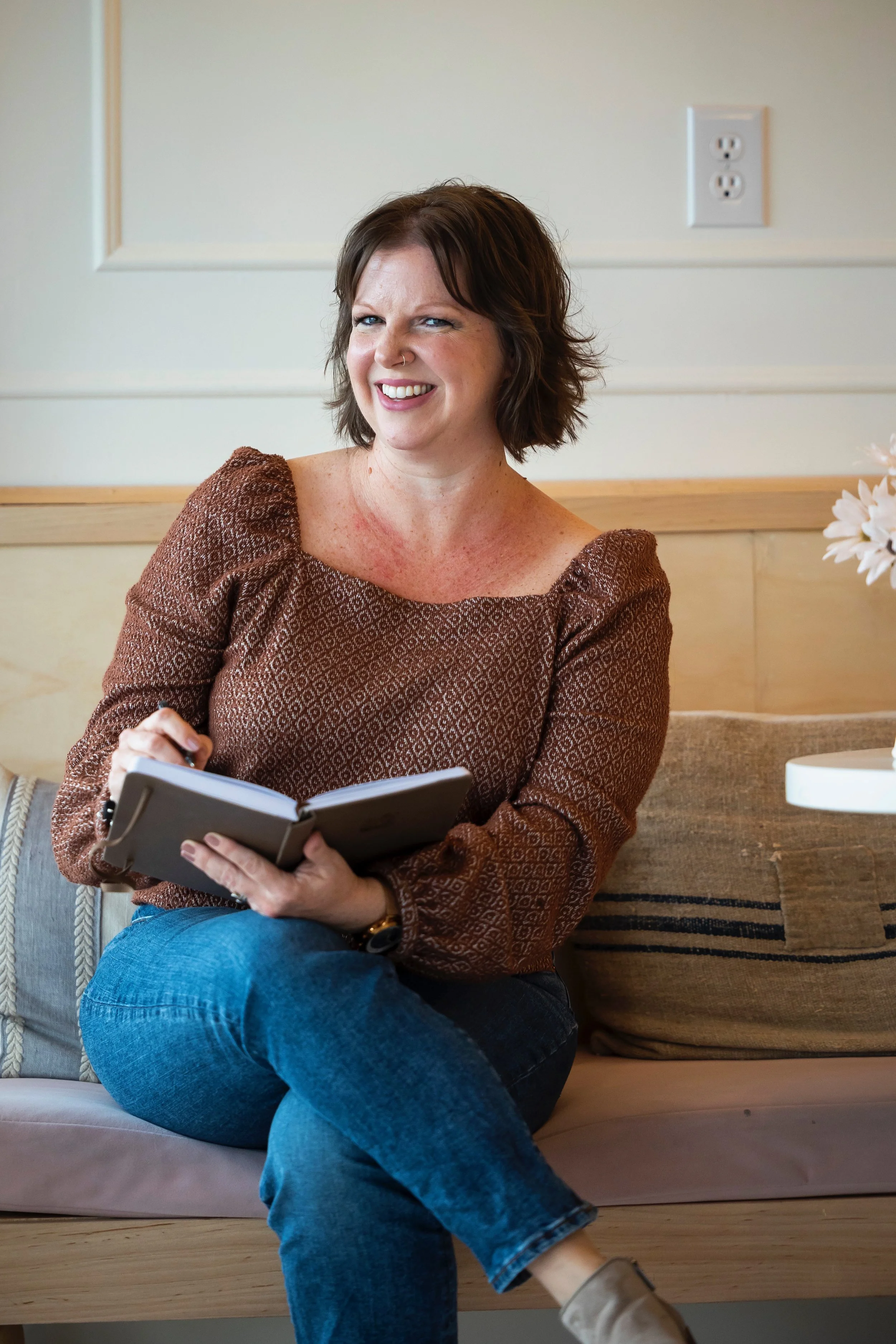 A woman with short brown hair, wearing a brown patterned top, sitting on a beige couch with a notebook, smiling and looking at the camera.