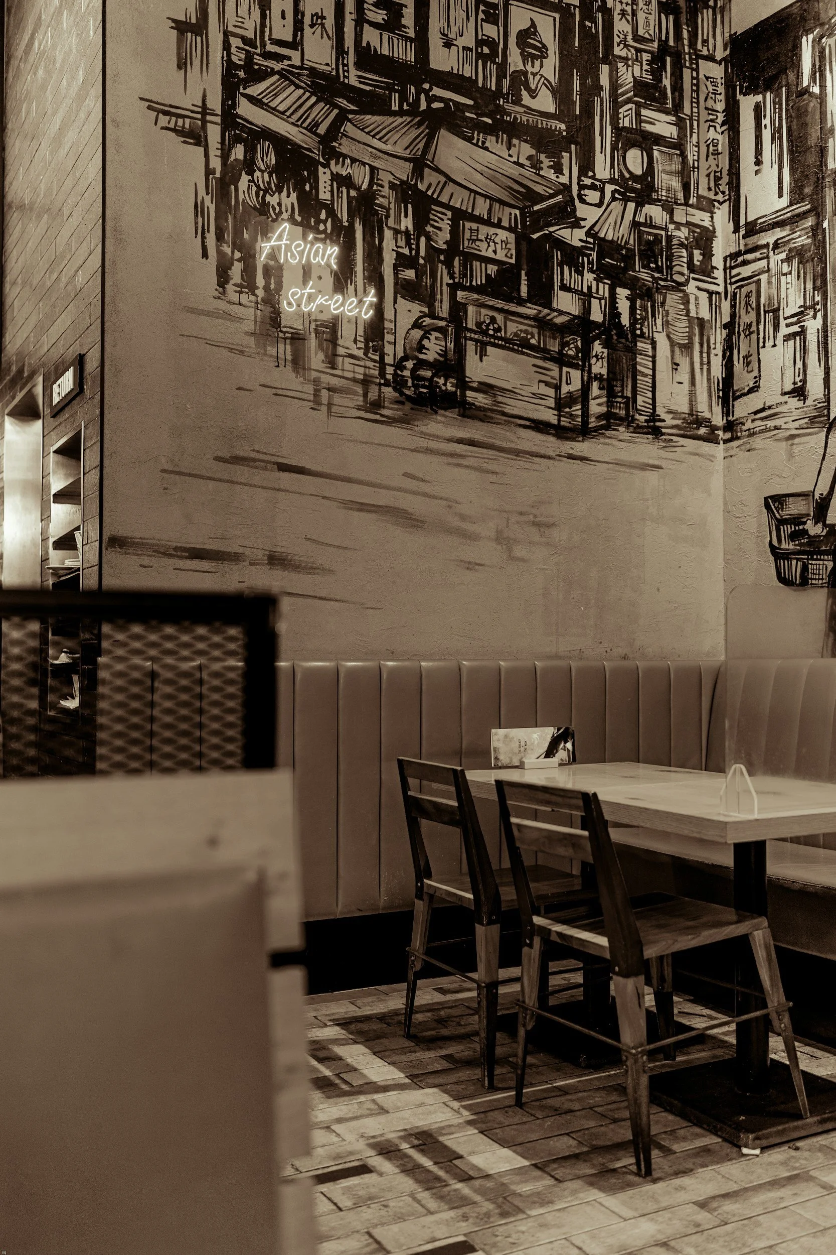 Interior of a restaurant with a large wall mural depicting an Asian street scene in black and white. The mural features buildings with signs in Chinese characters and a prominent neon sign that reads "Asian street." There are wooden chairs and tables in the foreground, and the floor has a tile pattern.
