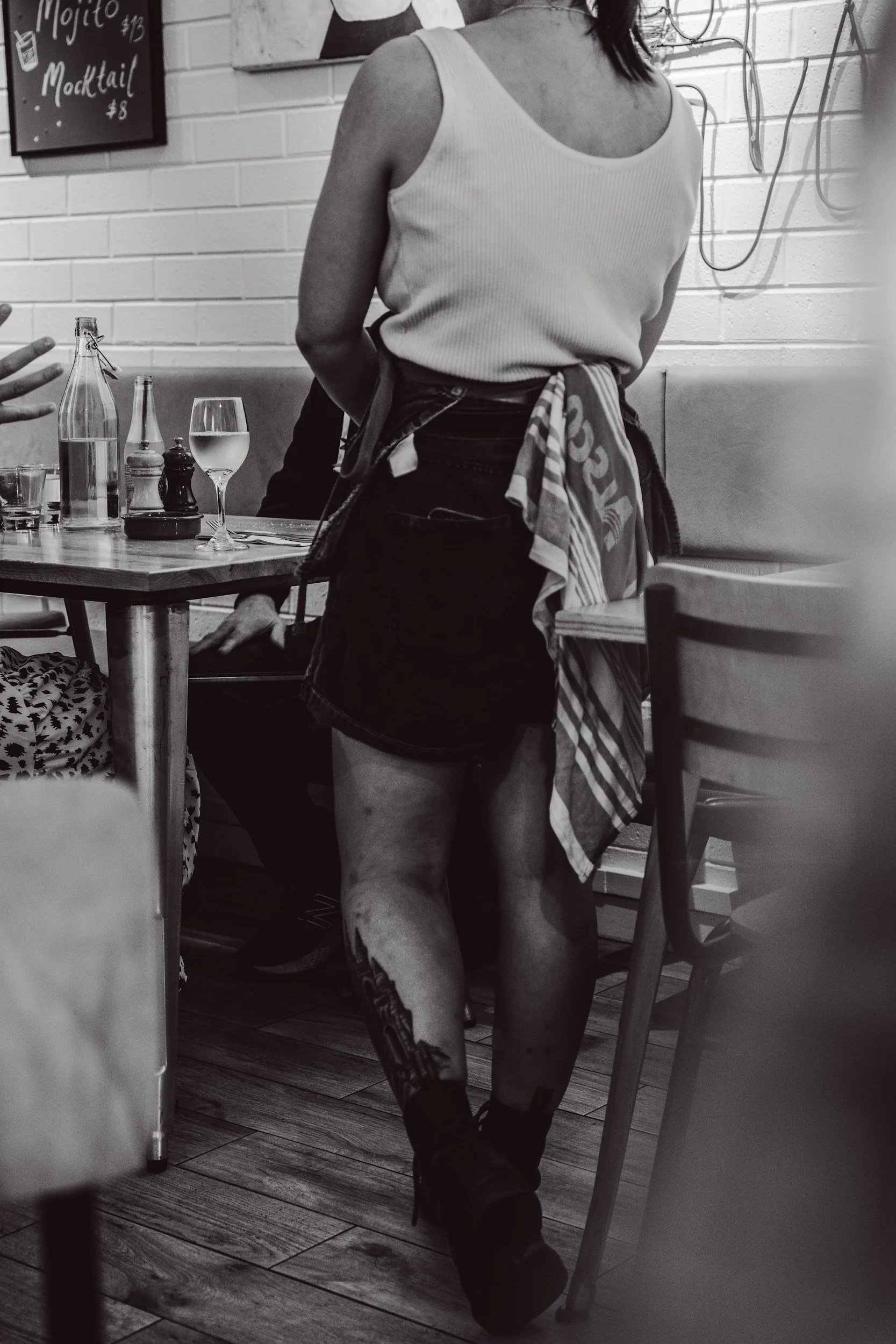 Person in a white tank top and black shorts standing in a restaurant, with a tattoo on their left leg, surrounded by table settings including glasses and bottles, and a brick wall with a chalkboard menu in the background.
