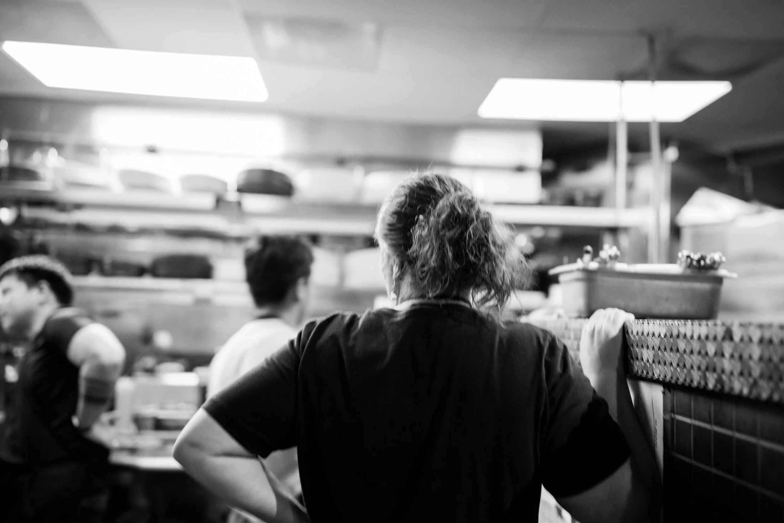 Black and white photo of a woman with curly hair seated in an indoor setting, surrounded by other people, with bright ceiling lights and shelves in the background.
