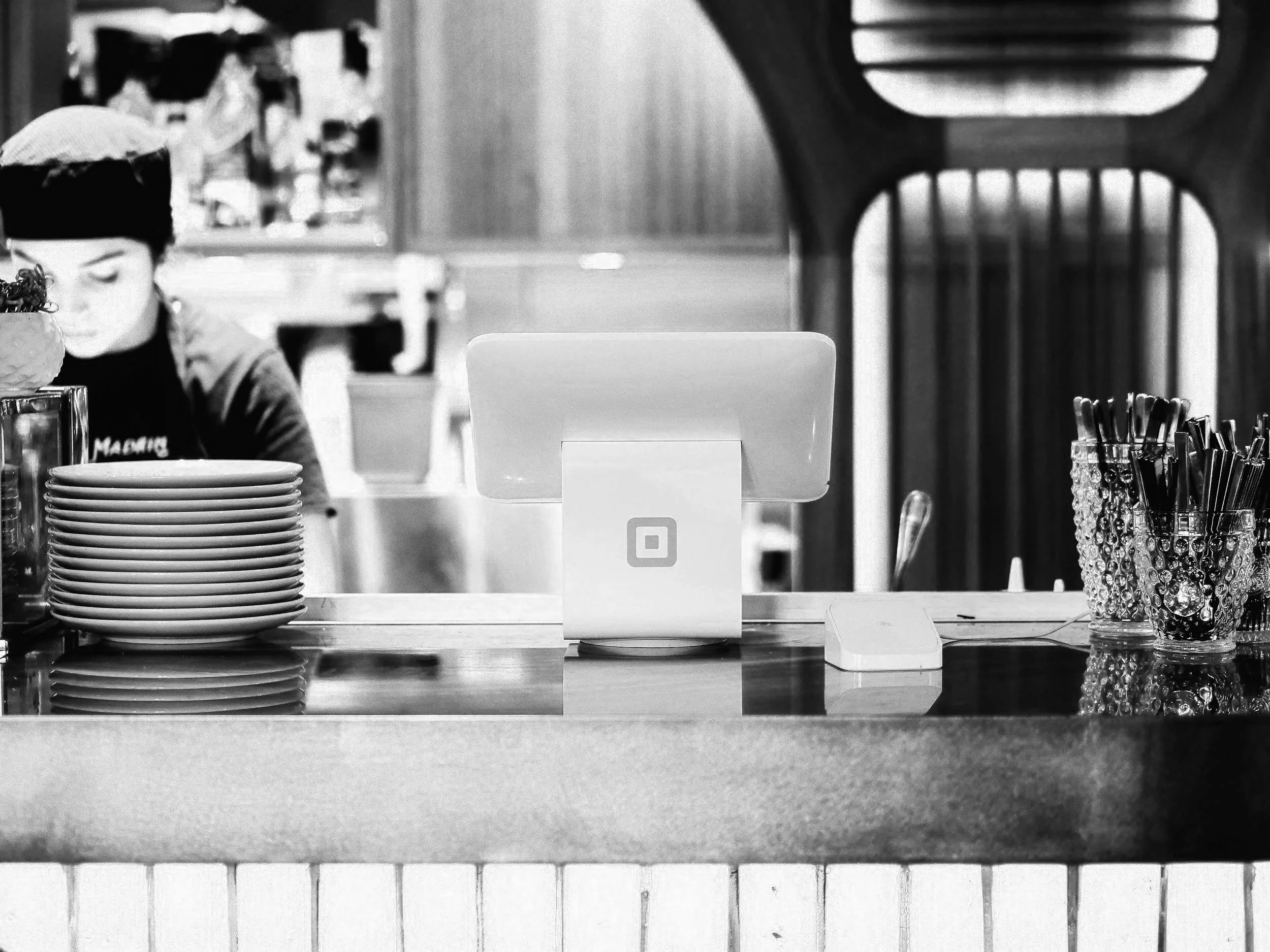 A black and white photo of a restaurant or cafe counter with stacked plates on the left, a computer or register in the center, and pens or straws in a container on the right. A person wearing a hat is partially visible behind the counter.