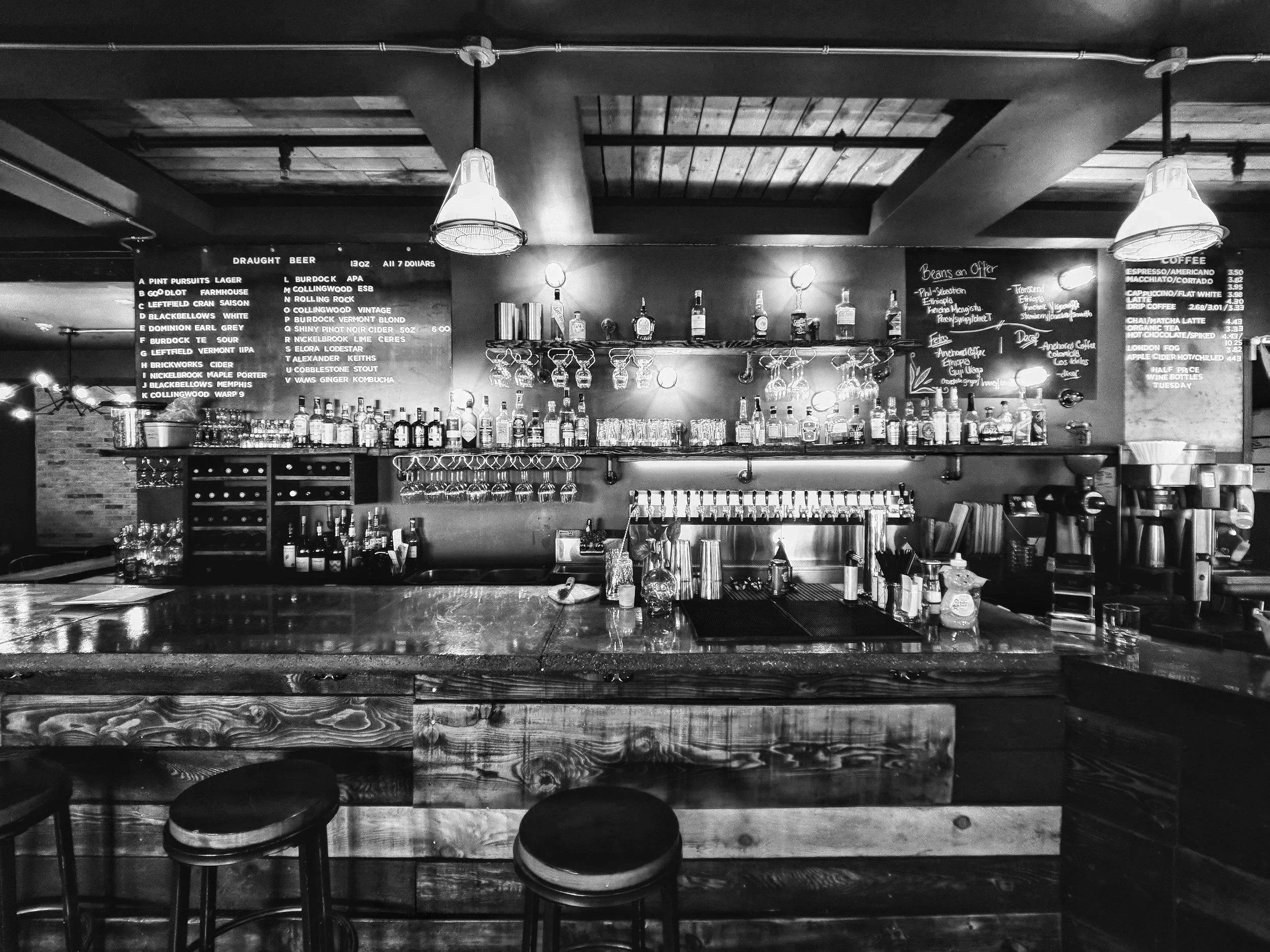 A black-and-white photo of a bar interior with a counter, shelves of bottles, hanging glasses, and chalkboard menus listing drinks and prices.