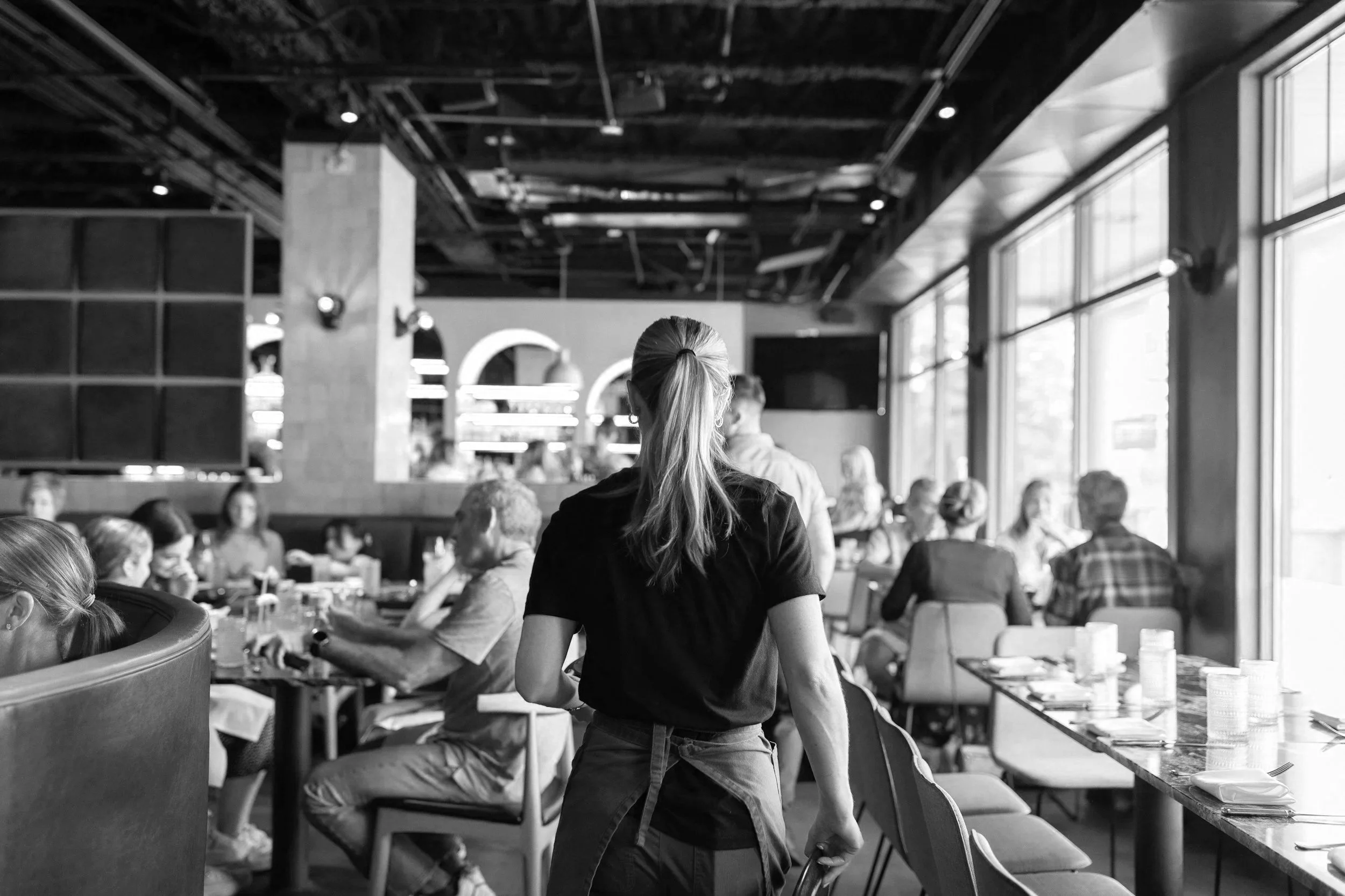 A woman with a ponytail wearing a black t-shirt stands in a busy restaurant, facing away from the camera, with other patrons sitting at tables and food on the tables.