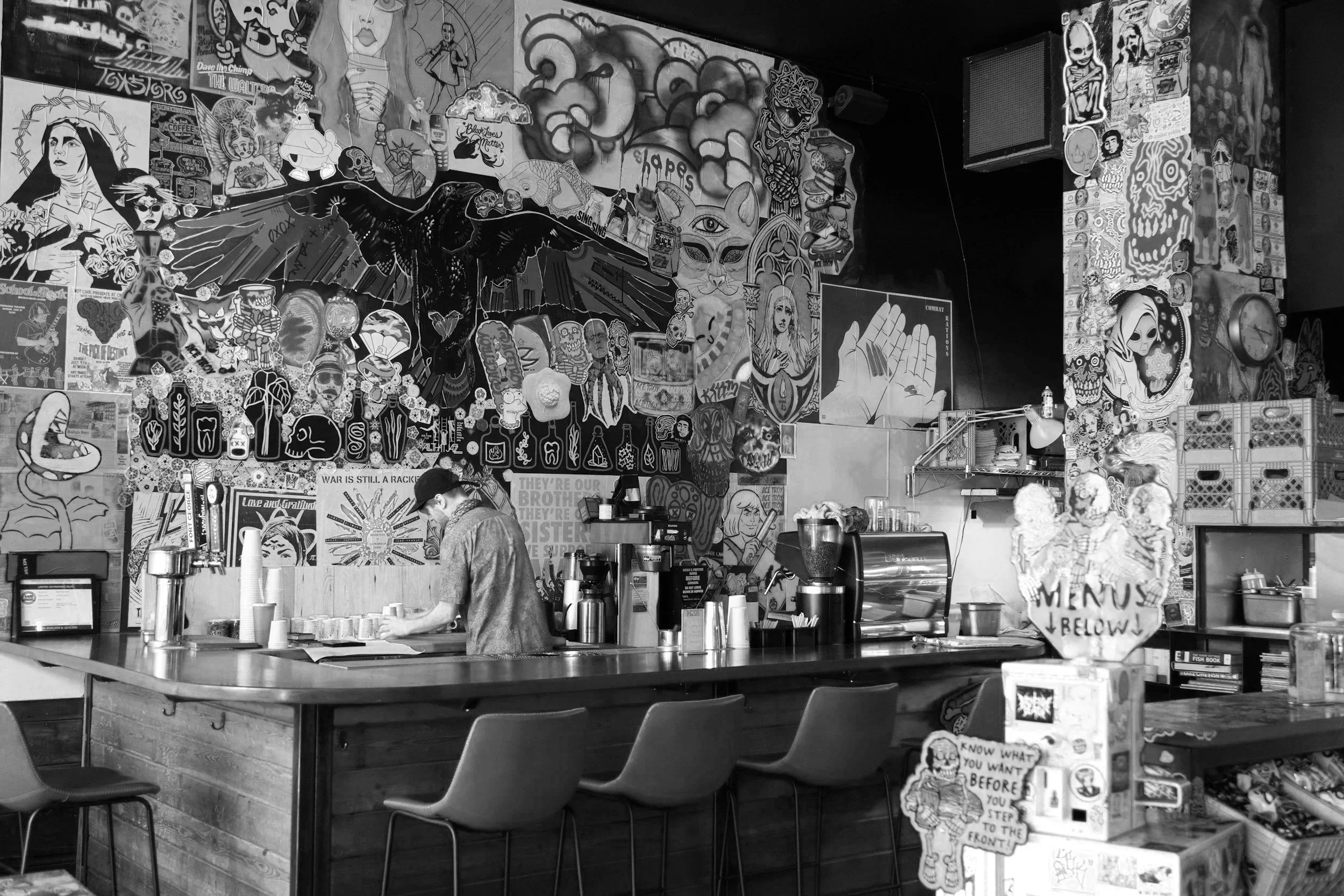 A black and white photo of a cafe or coffee shop with numerous posters and artwork on the walls. A person wearing a cap and apron is working behind the counter, preparing beverages. There are several cups, a coffee machine, and menu items visible on the counter, along with chairs and tables in the foreground.