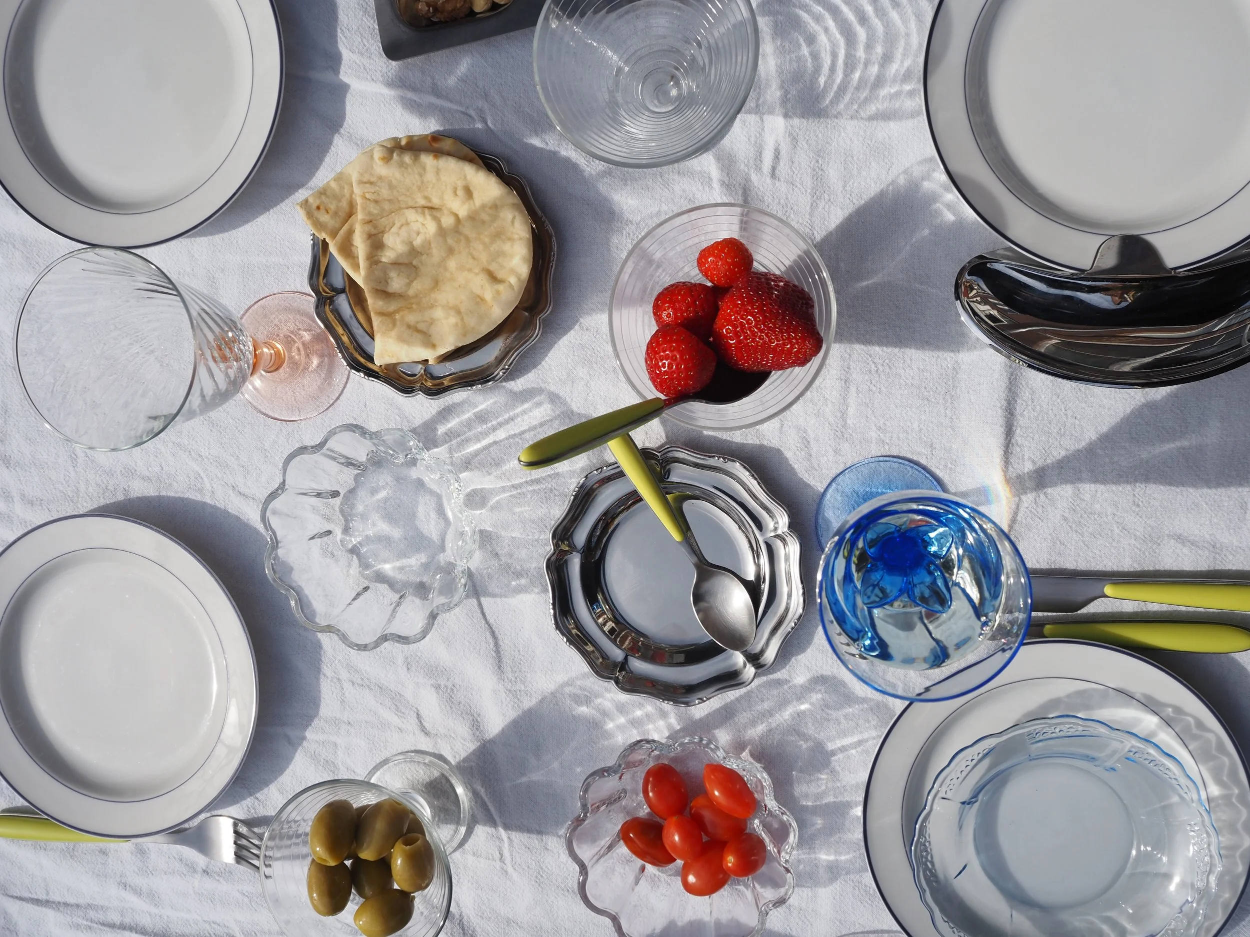 A top-down view of a table with various dishes and glasses, including a bowl of strawberries, an empty plate, a bowl of cherry tomatoes, a bowl of green olives, a bowl of flatbread, and several empty and partially filled wine glasses on a white tablecloth.